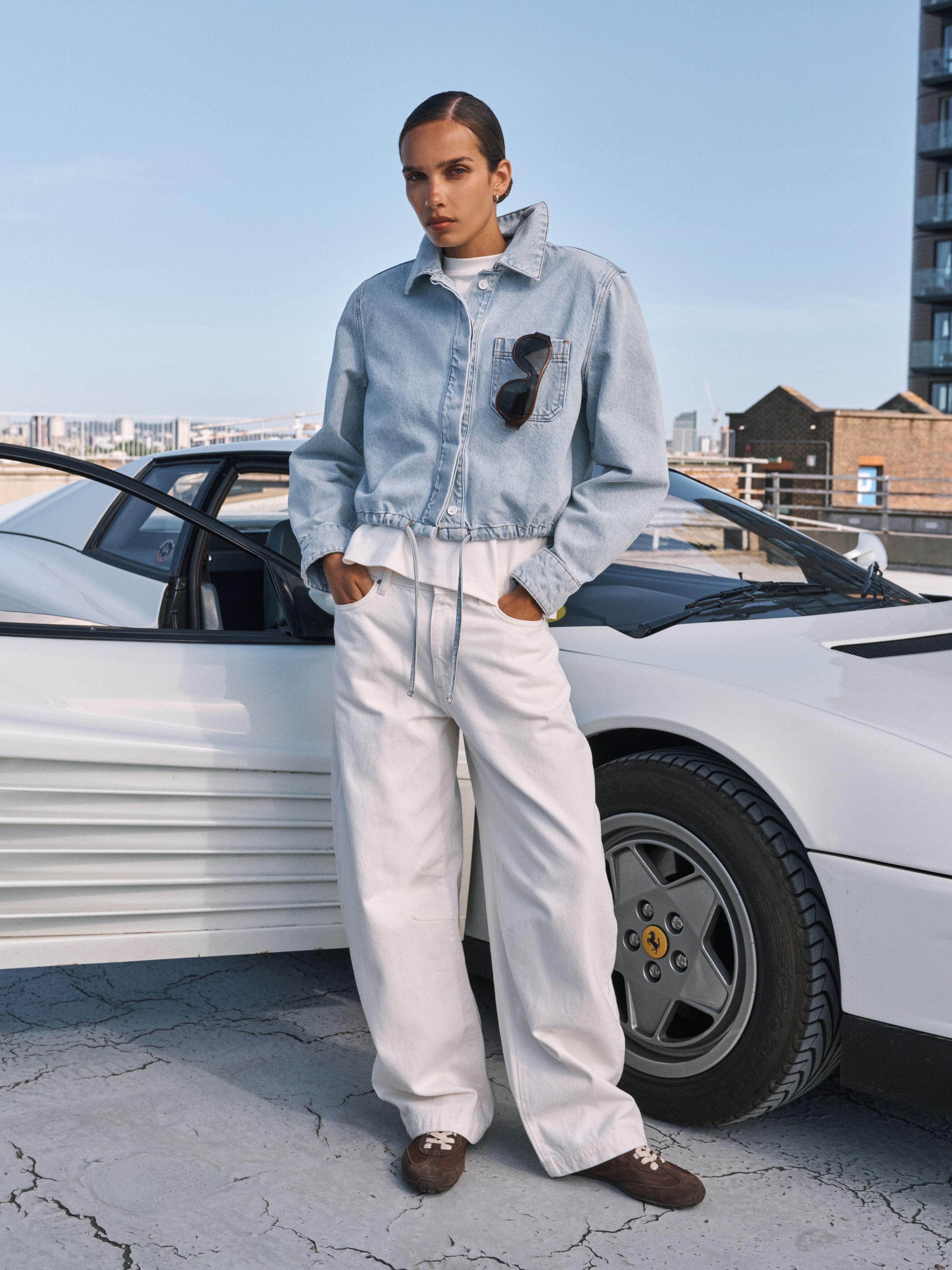 A model stands outdoors next to a white sports car, wearing a light-wash denim cropped jacket, white wide-leg trousers, and brown loafers. Sunglasses hang from the collar of the jacket.