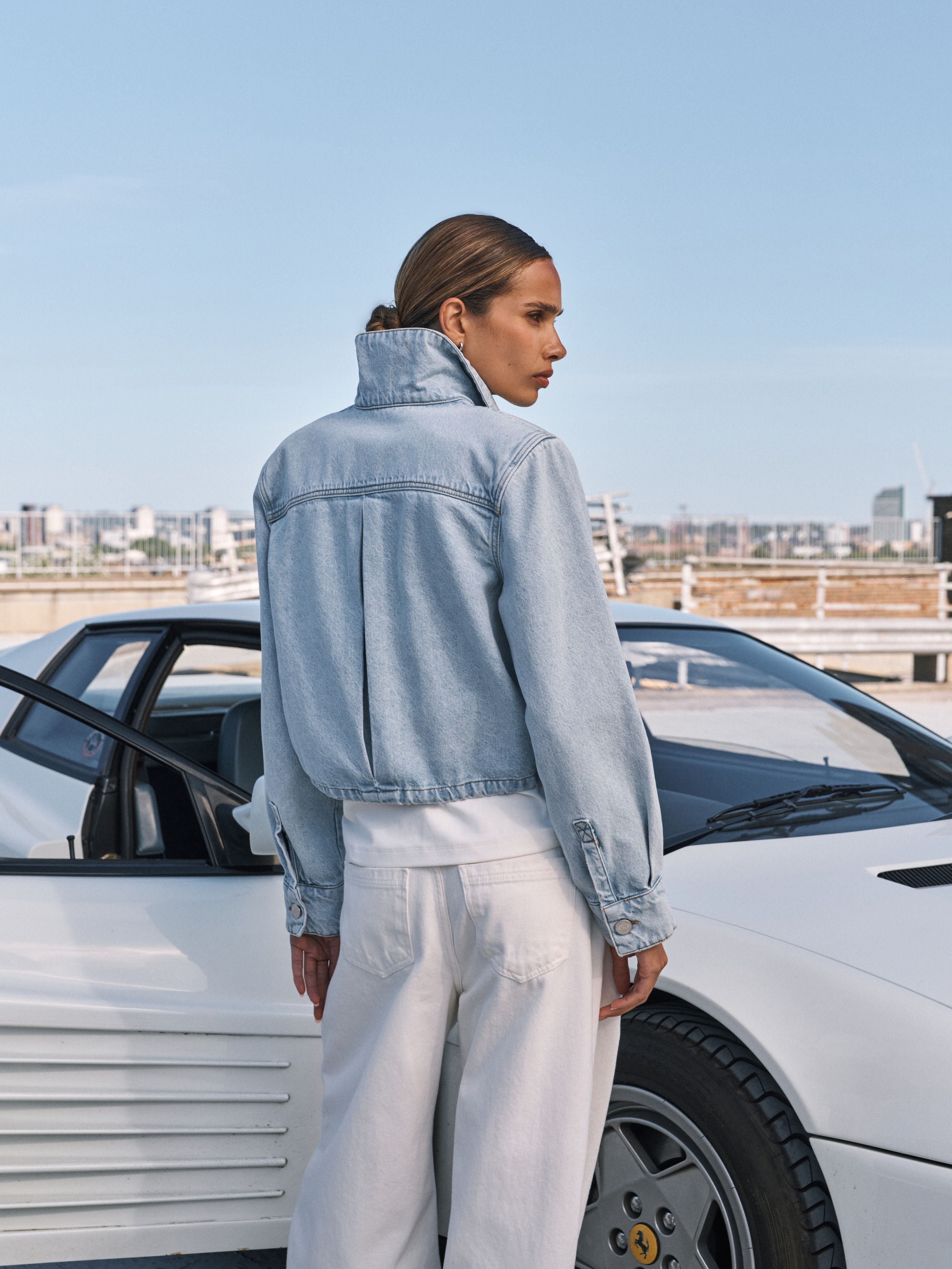 A woman stands outdoors in front of a white sports car, shown from the back and side. She is wearing a light-wash denim cropped jacket with a high collar and white wide-leg pants.