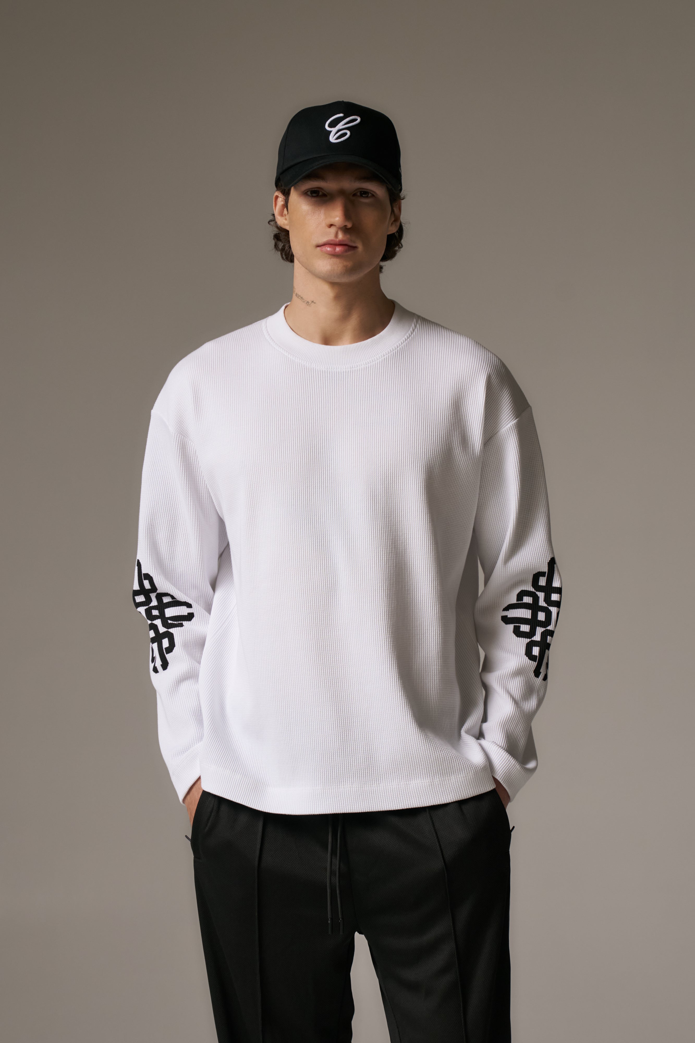Person wearing a white long sleeved t-shirt with black patterns and a black cap on a grey background