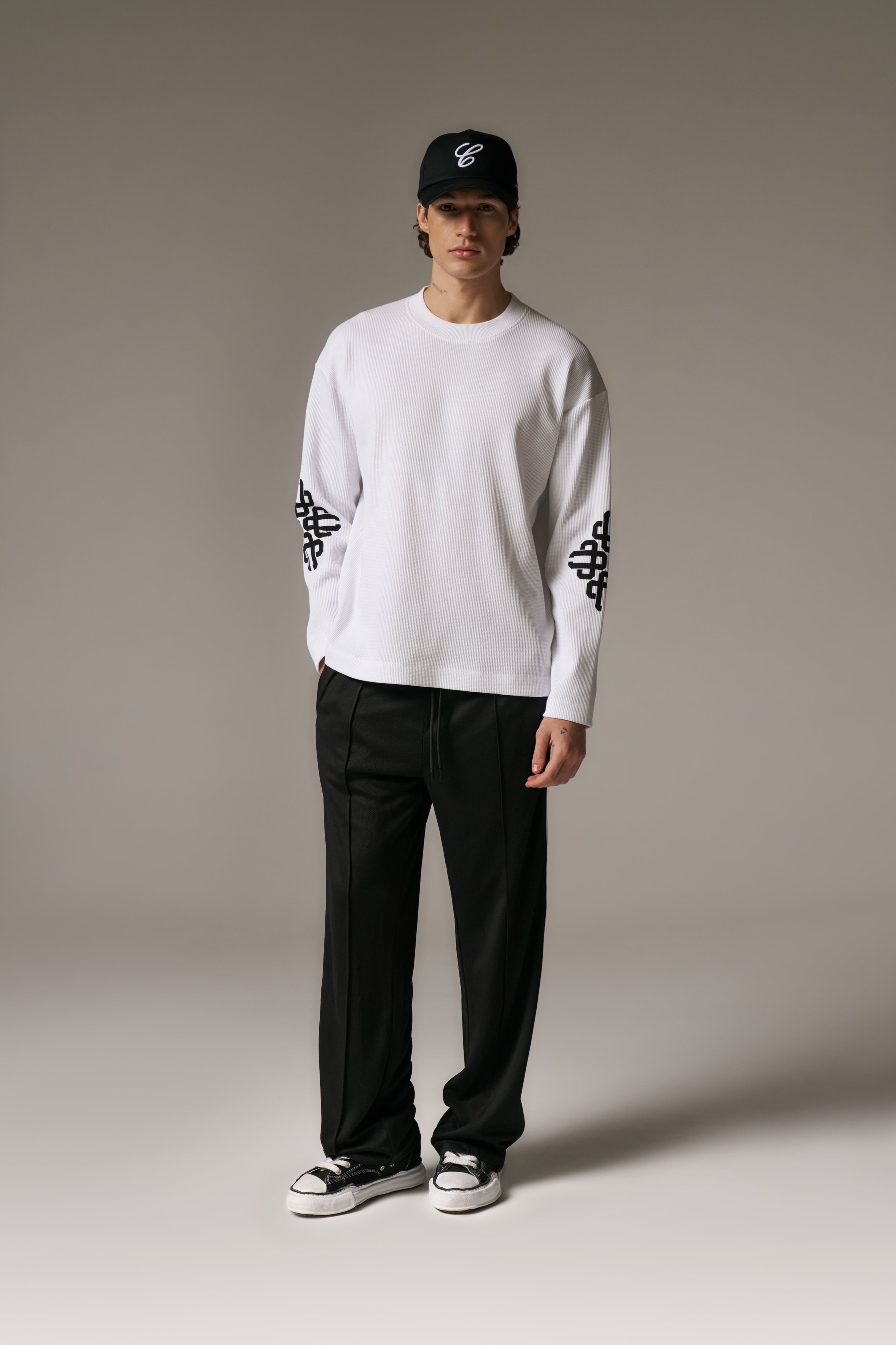 Person wearing a white long sleeved t-shirt with black patterns and black pants on a plain background