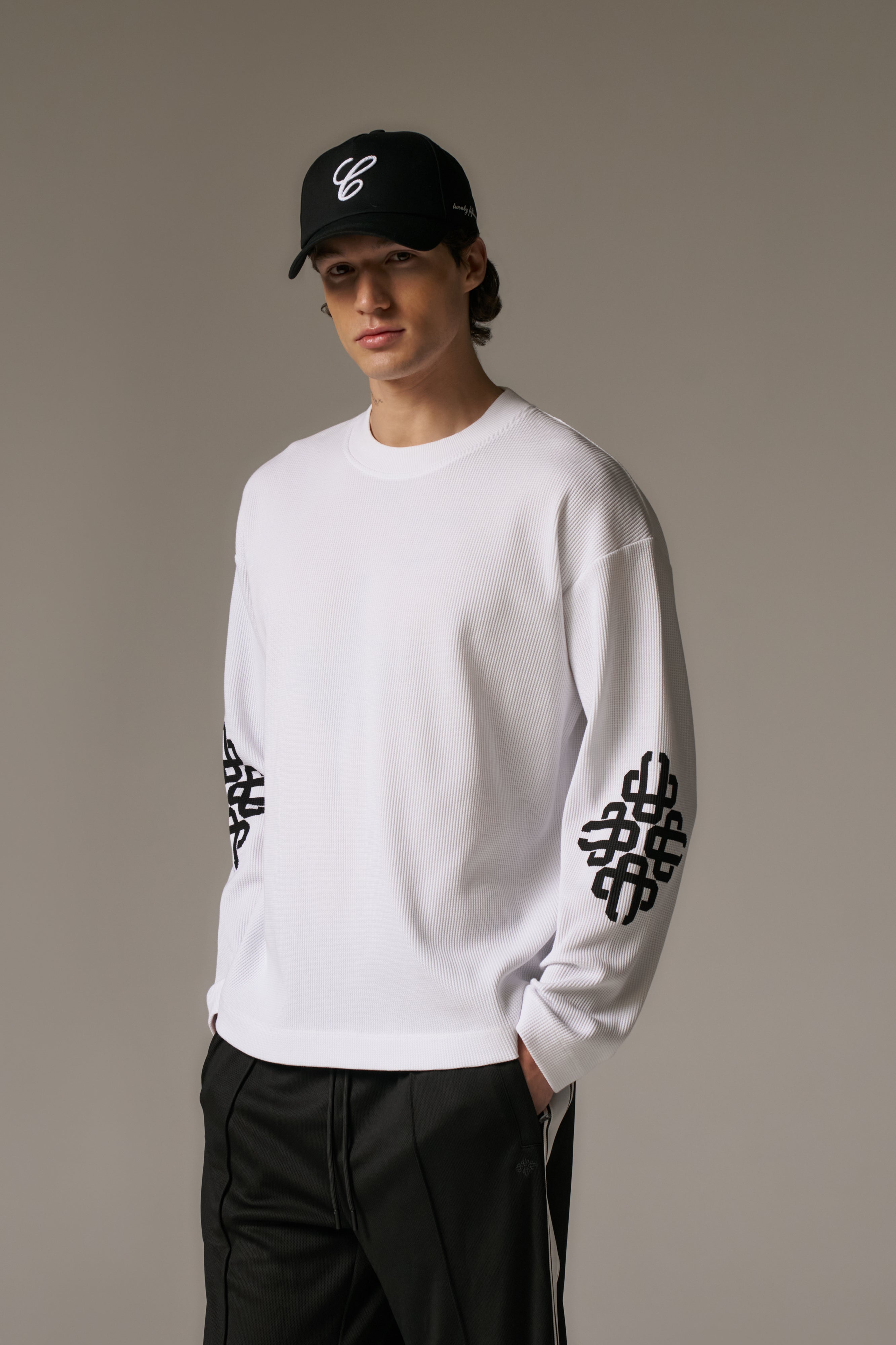 Person wearing a white long sleeved t-shirt with black patterns on sleeves and a black cap against a neutral background