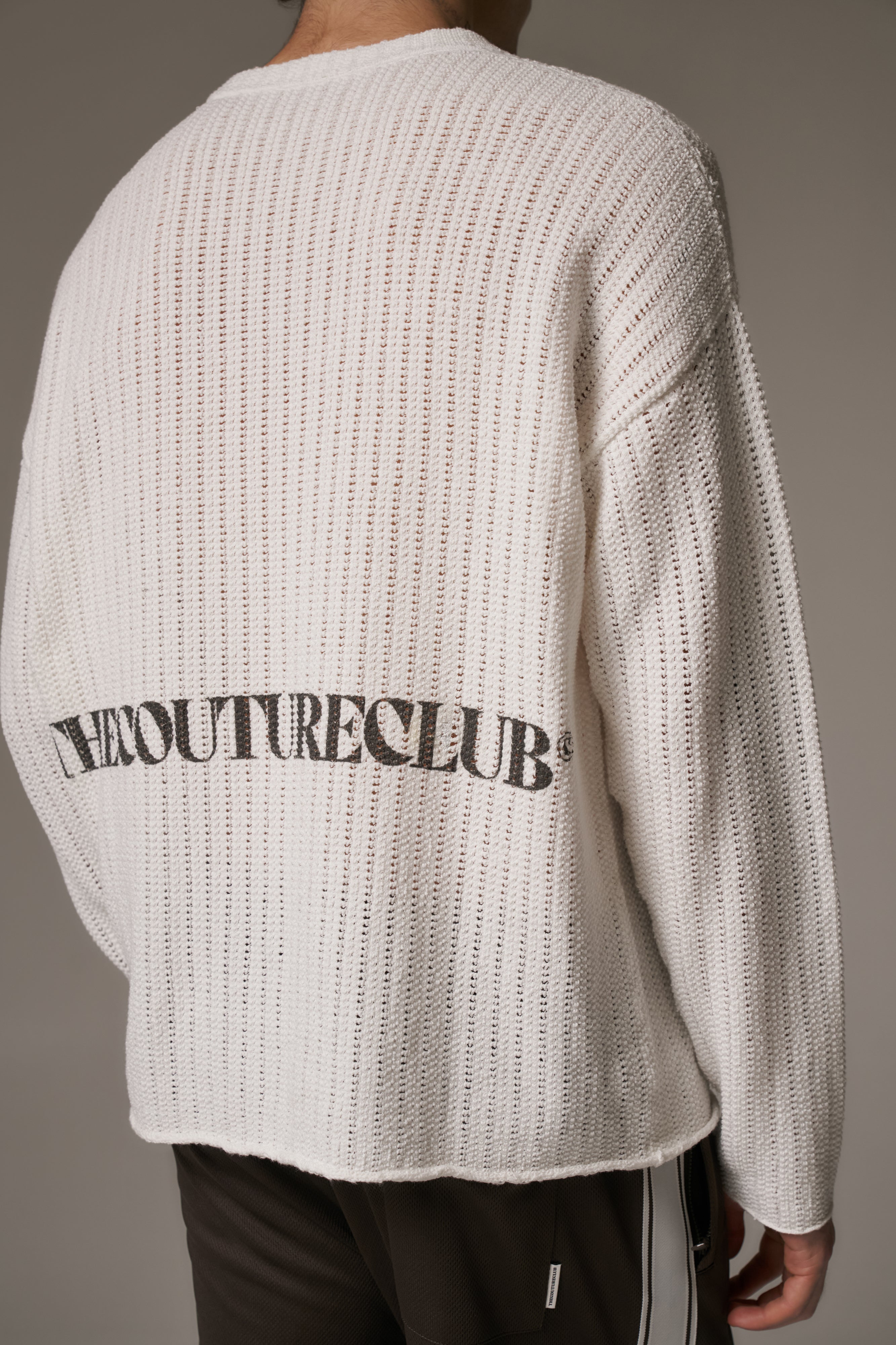 Person wearing a white crochet knit long sleeved t-shirt with branded text on the back against a neutral background.