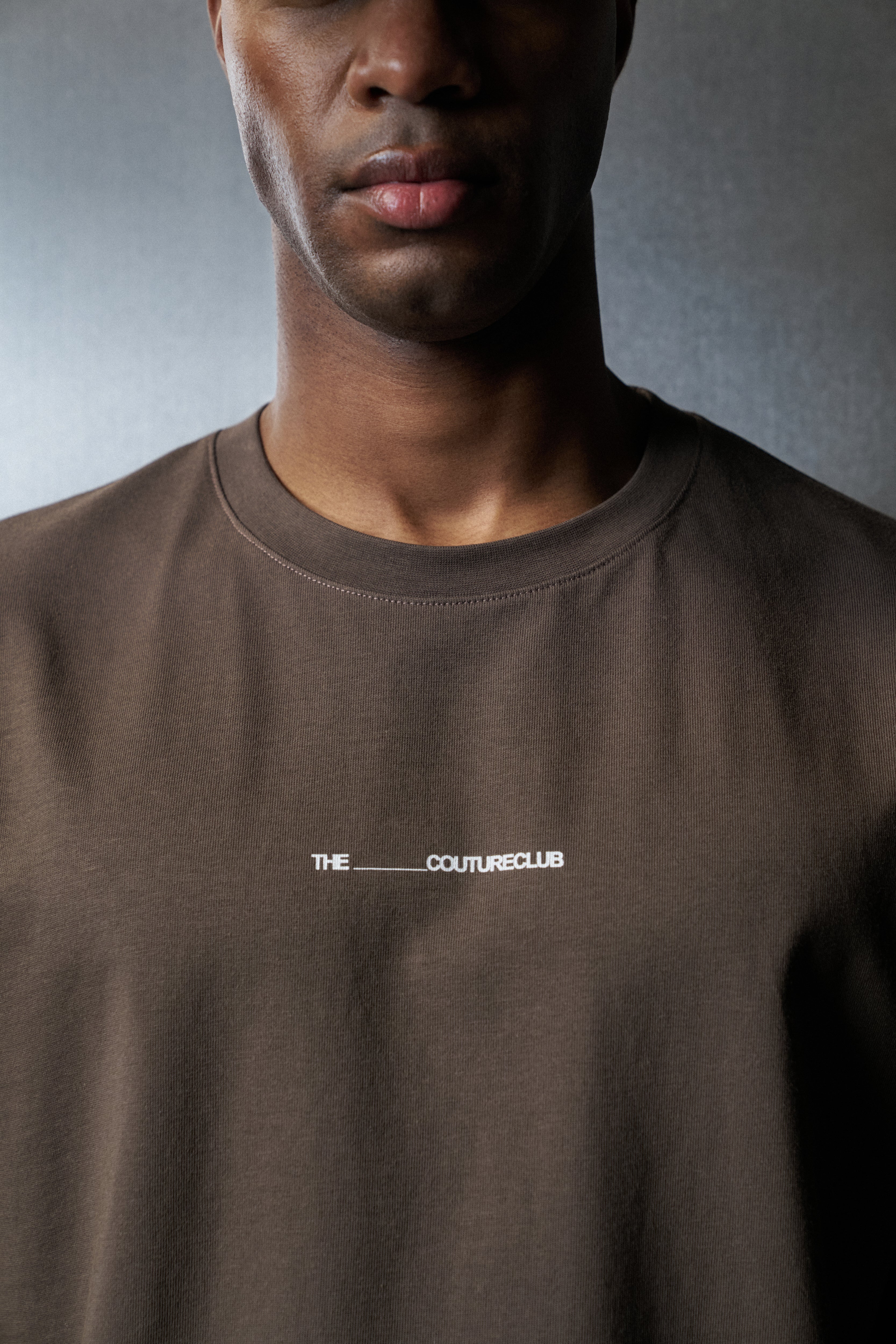 Man wearing a brown t-shirt with 'The Couture Club' logo on a grey background