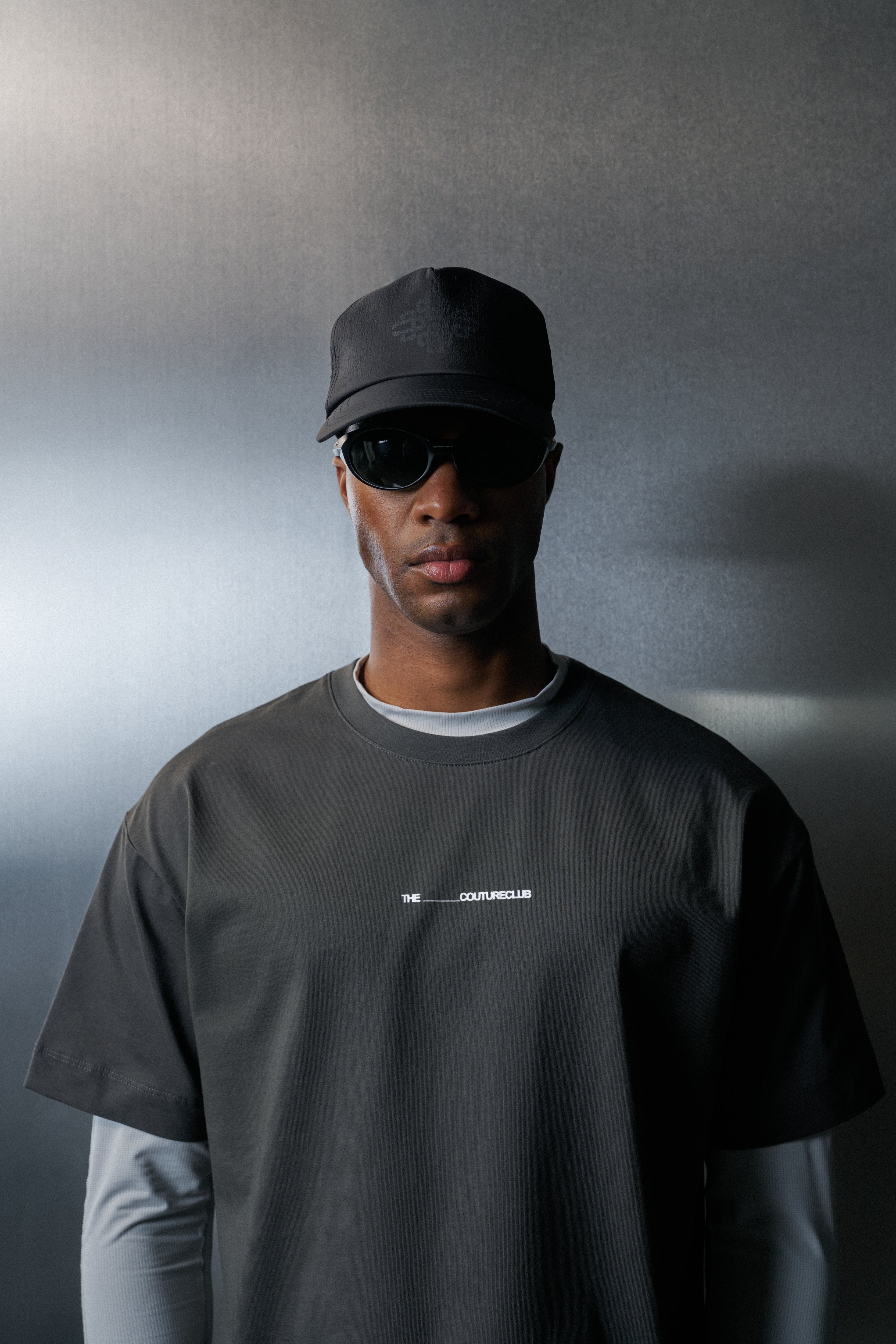 Man wearing a black cap and sunglasses with a dark t-shirt against a grey background