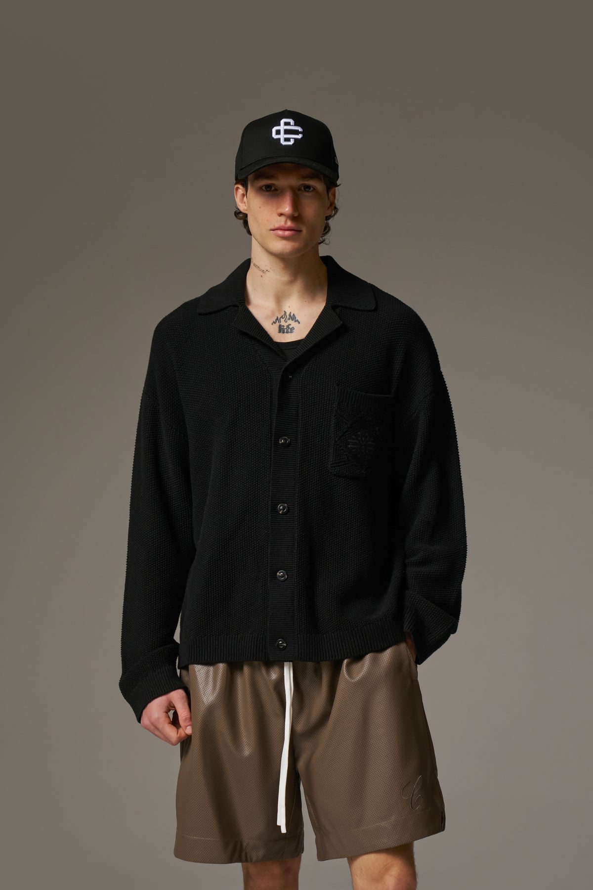 A person wearing a long-sleeved black shirt with a ribbed collar, crochet knit fabric, and an embroidered emblem on the chest pocket.