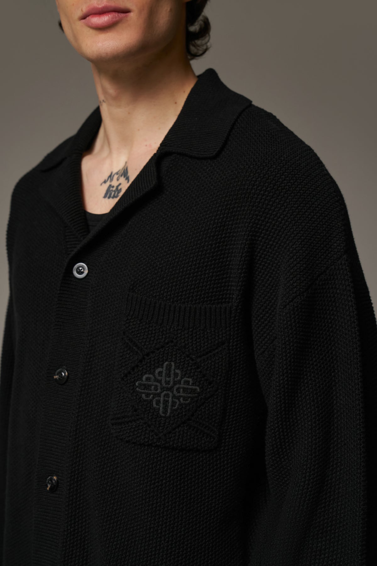 Close up detail shot of a person wearing a long-sleeved black shirt with a ribbed collar, crochet knit fabric, and an embroidered emblem on the chest pocket.
