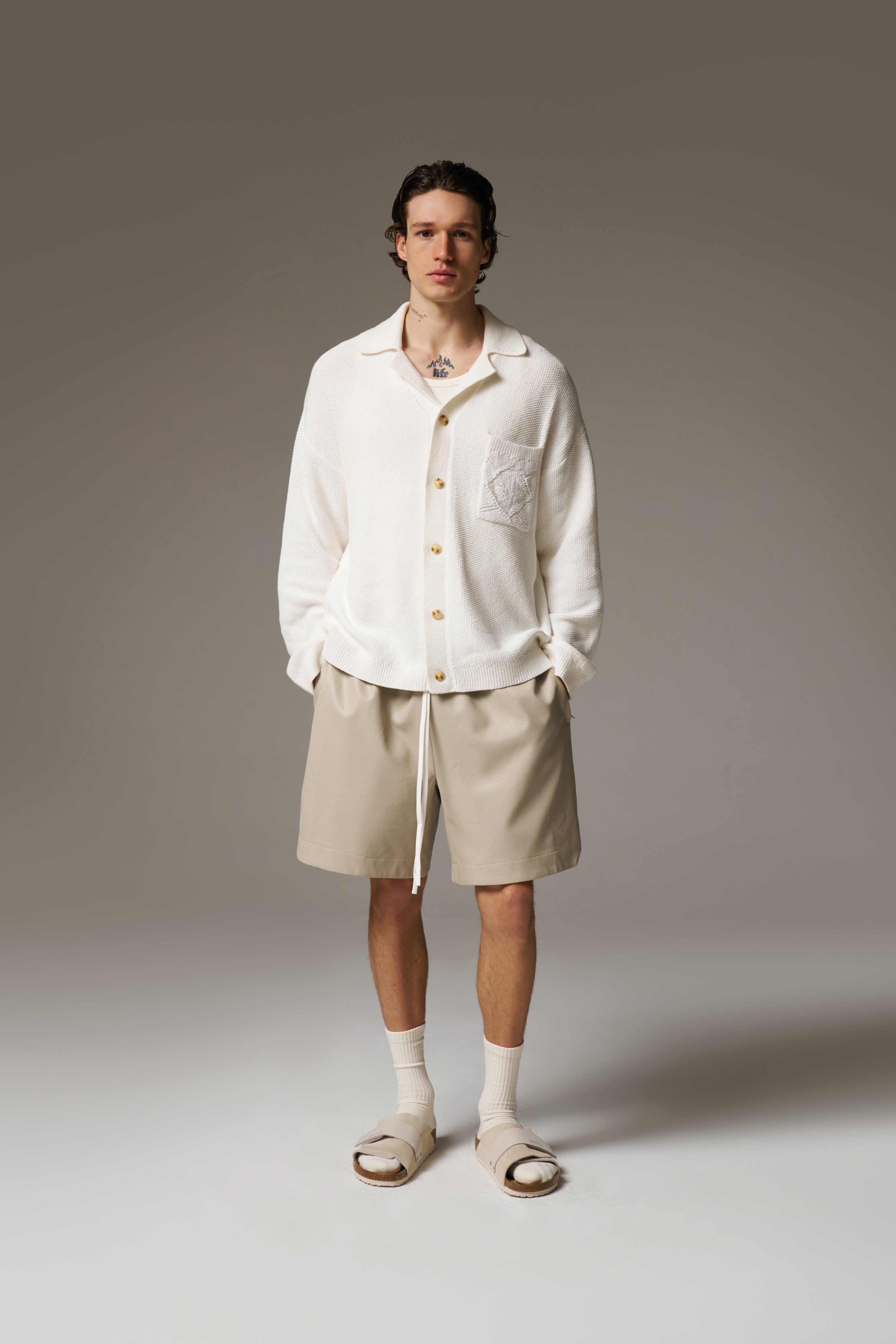 A man wearing a long sleeve crochet shirt and beige shorts against a neutral background.