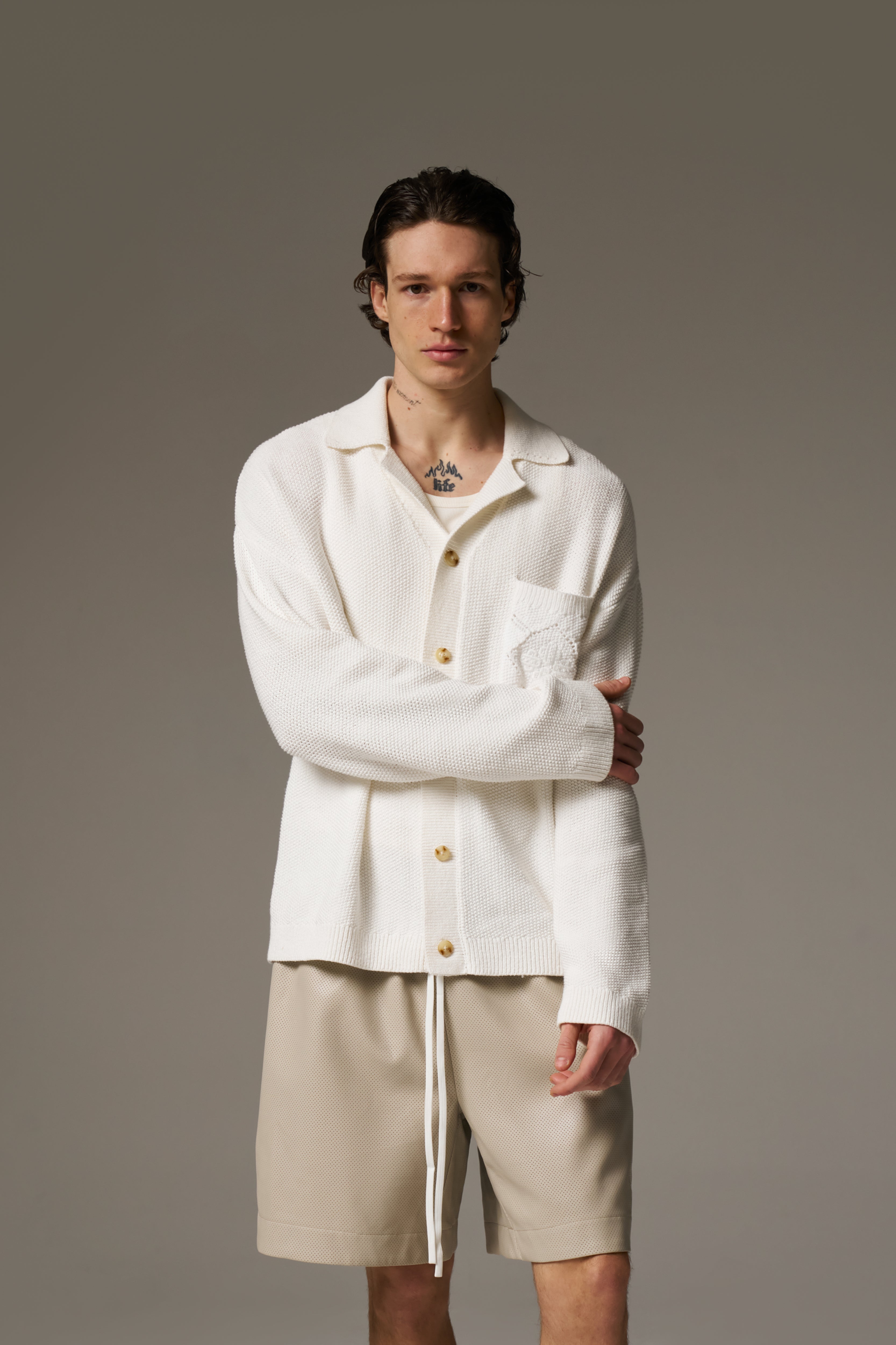 A man wearing a long sleeve crochet shirt and beige shorts against a neutral background.