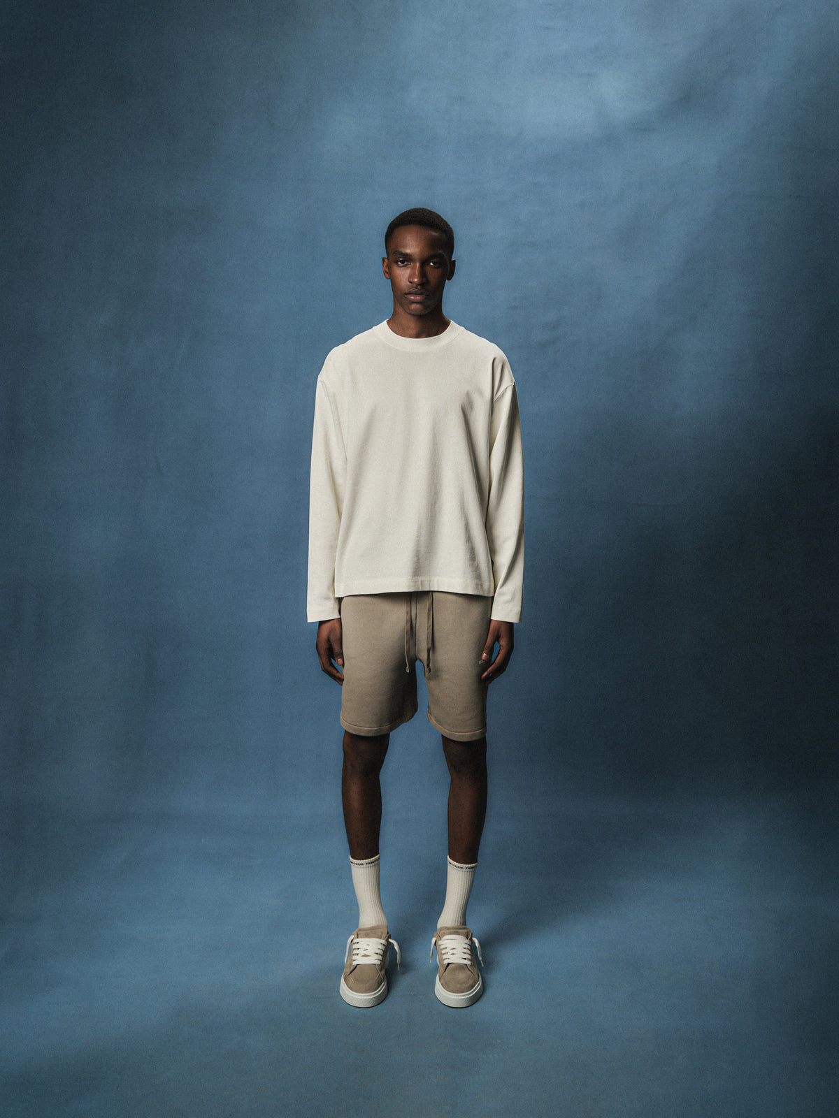 Person wearing an off white long sleeved t-shirt with beige shorts and shoes against a blue background