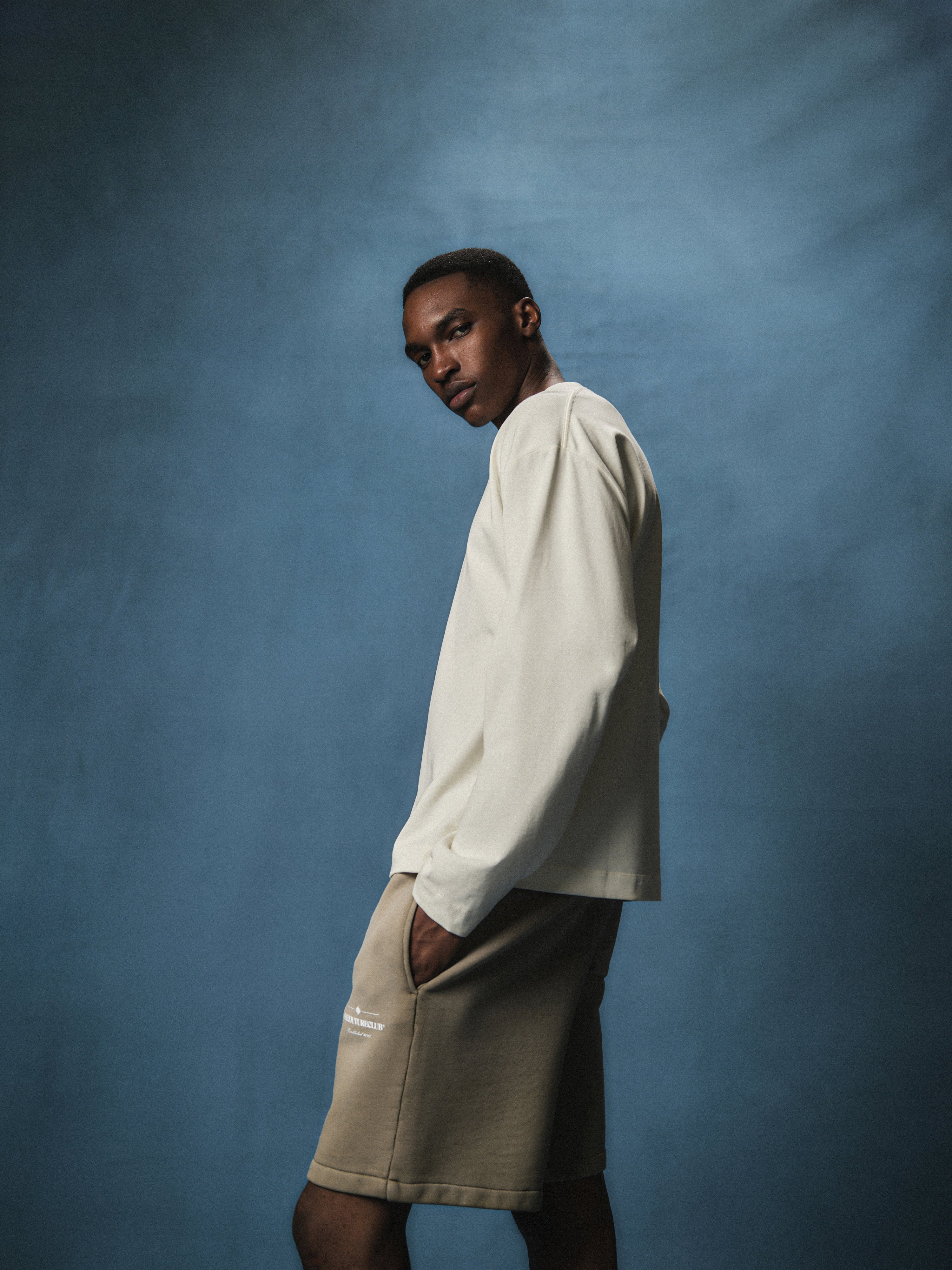 Side view of a man wearing an off white long sleeved t-shirt with beige shorts and shoes against a blue background
