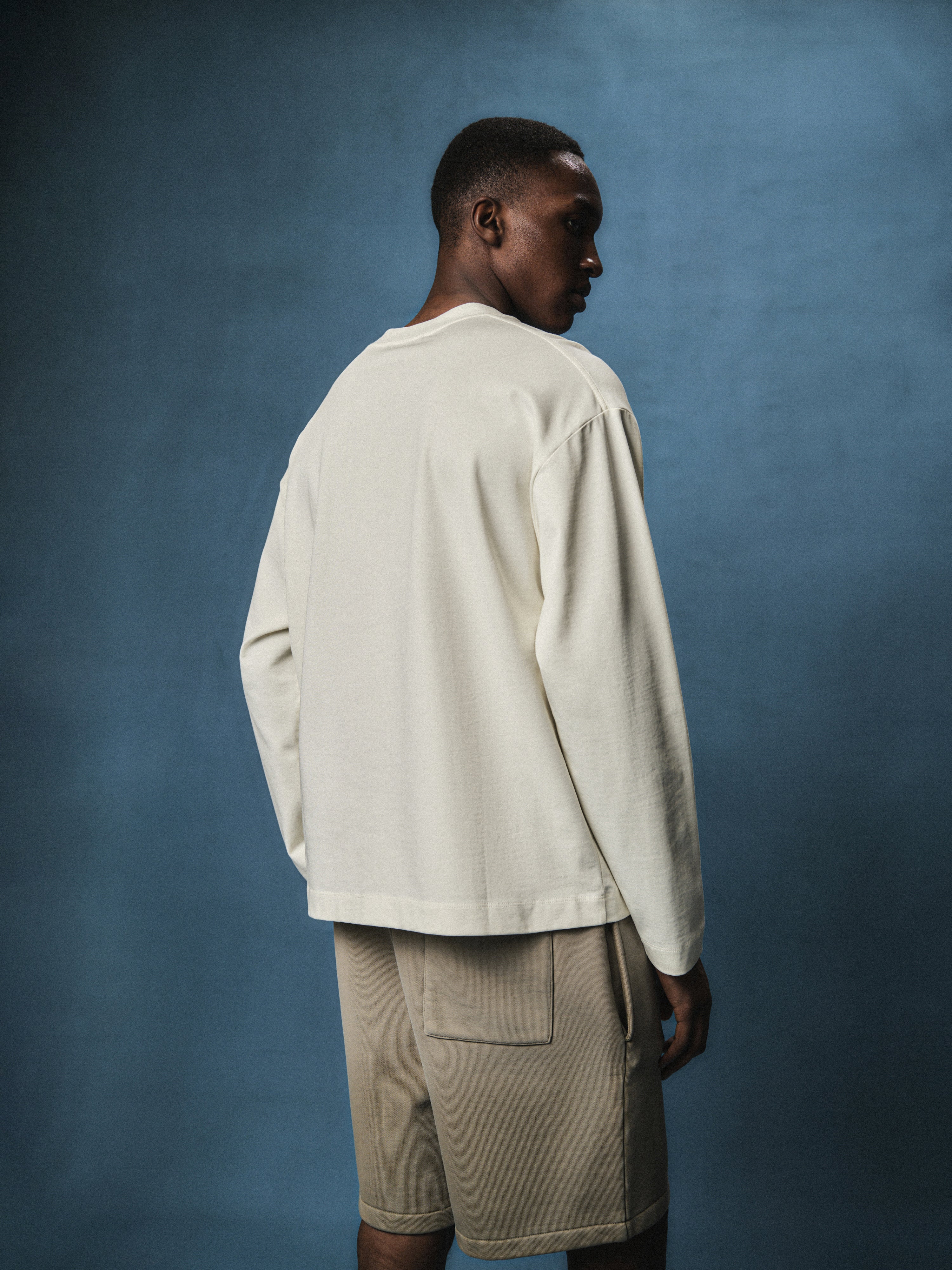 Back view of a man wearing a long sleeved off white t-shirt, wearing beige shorts with a dark blue back ground