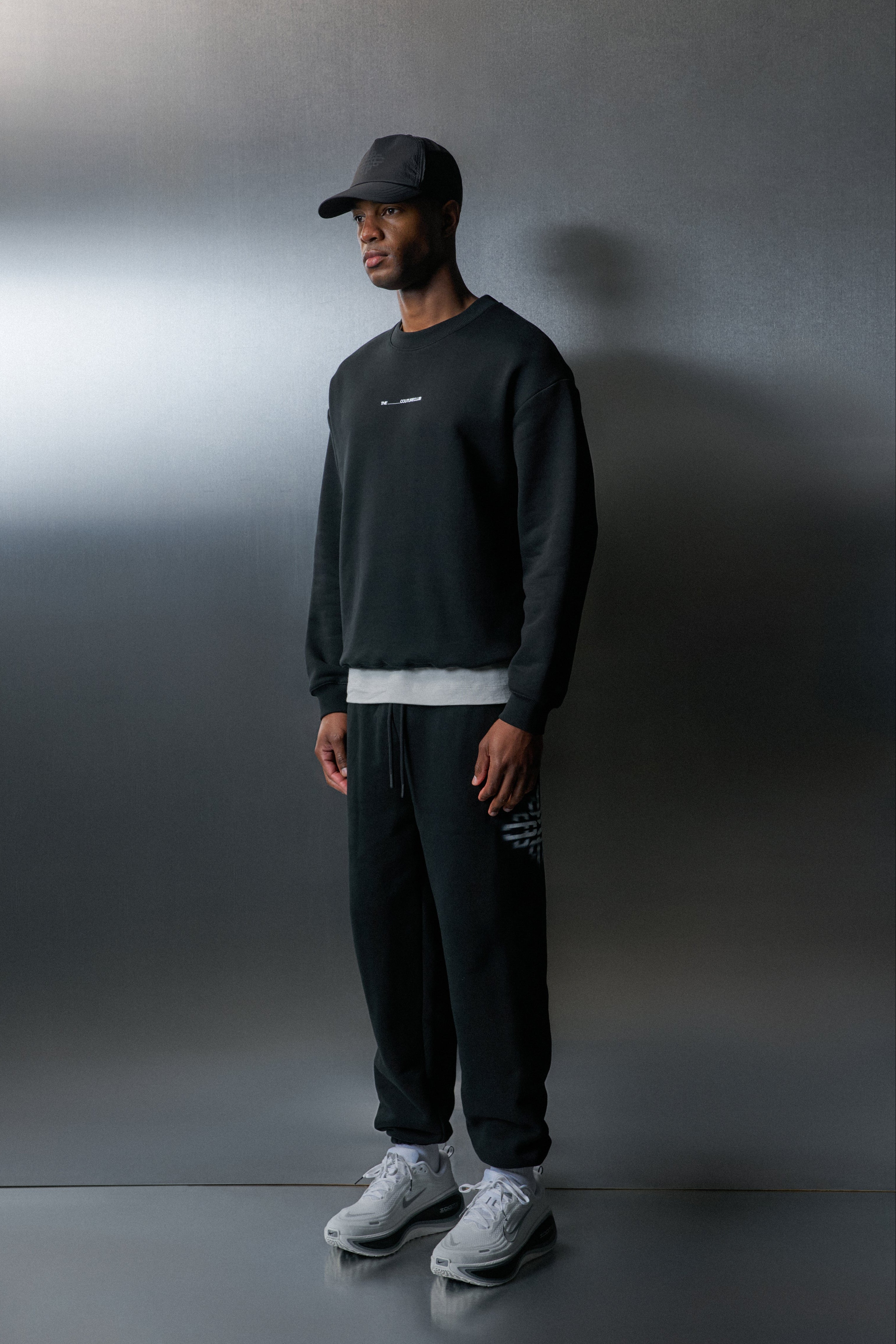 Person wearing a black sweatshirt, pants, and cap against a grey background