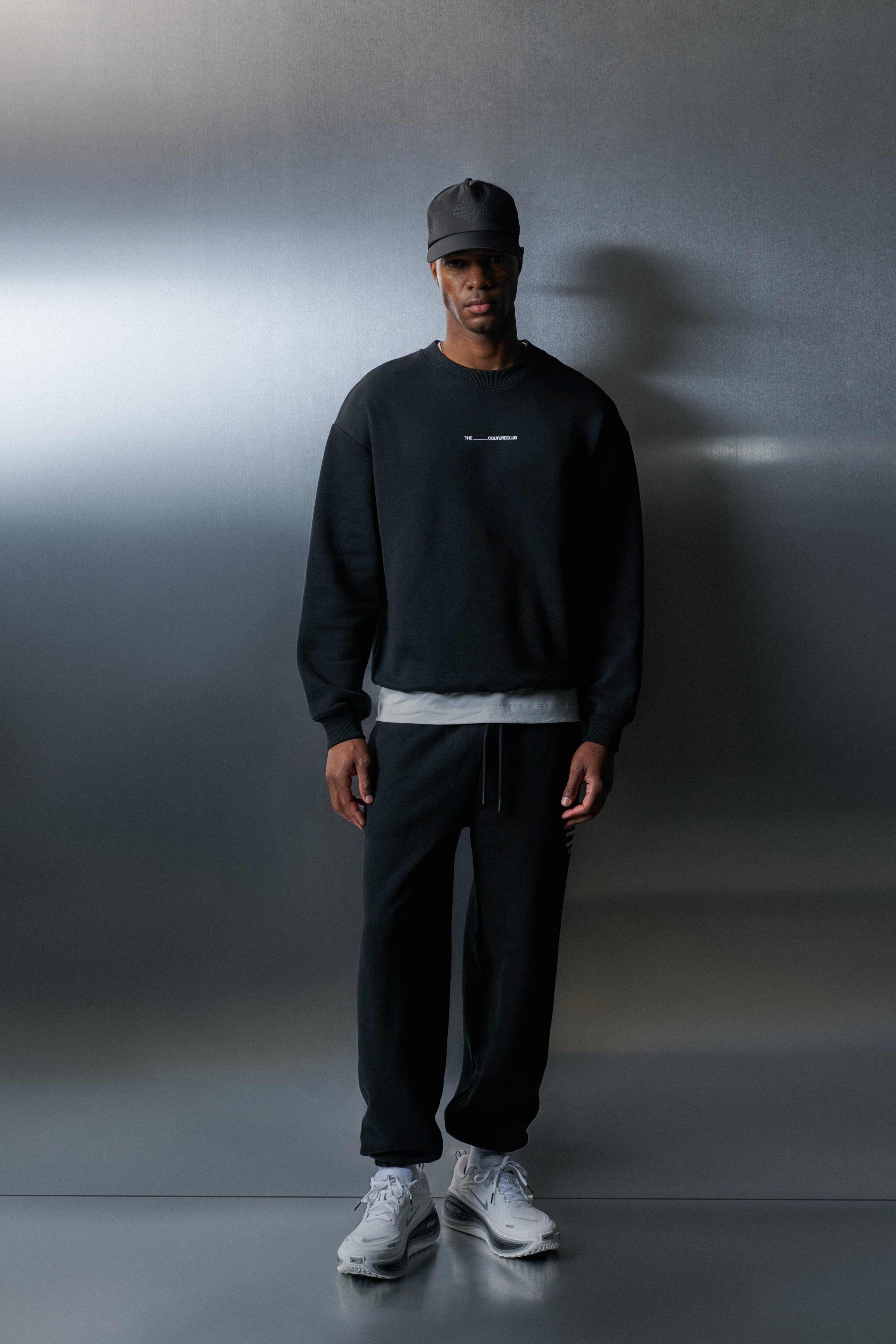 Person wearing a black sweatshirt, dark pants, and white sneakers against a grey background