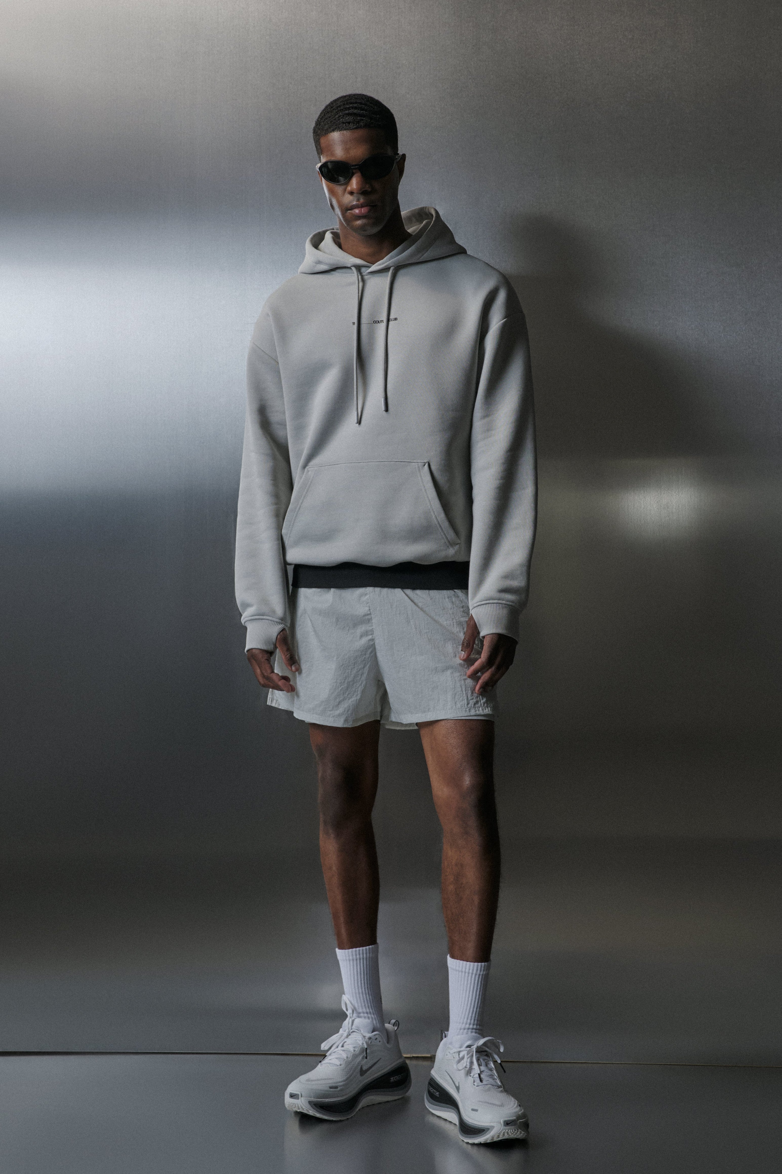 Man wearing a grey hoodie, shorts, and sneakers against a neutral background