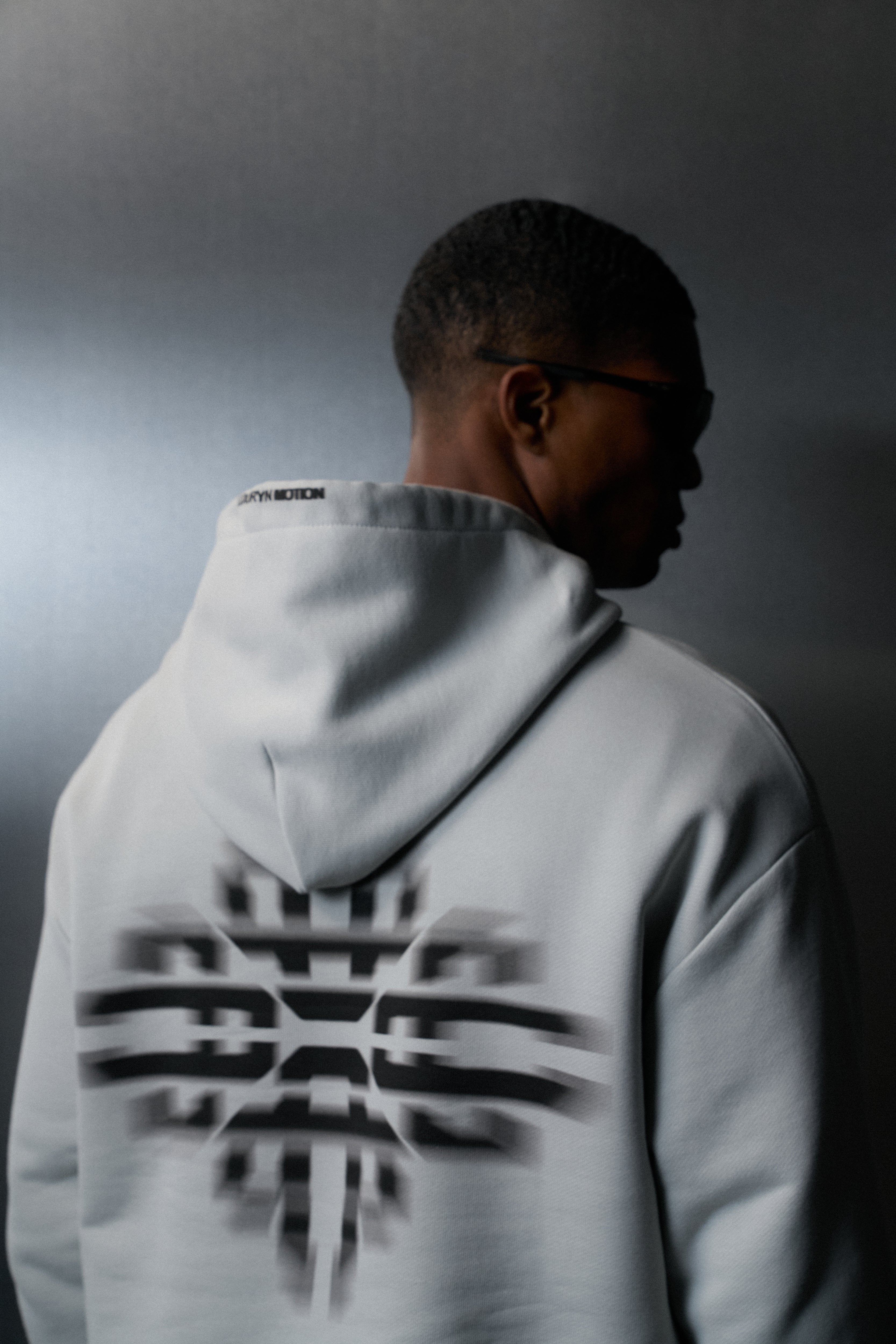 Person wearing a light grey hoodie with a logo on the back, standing against a dark background.