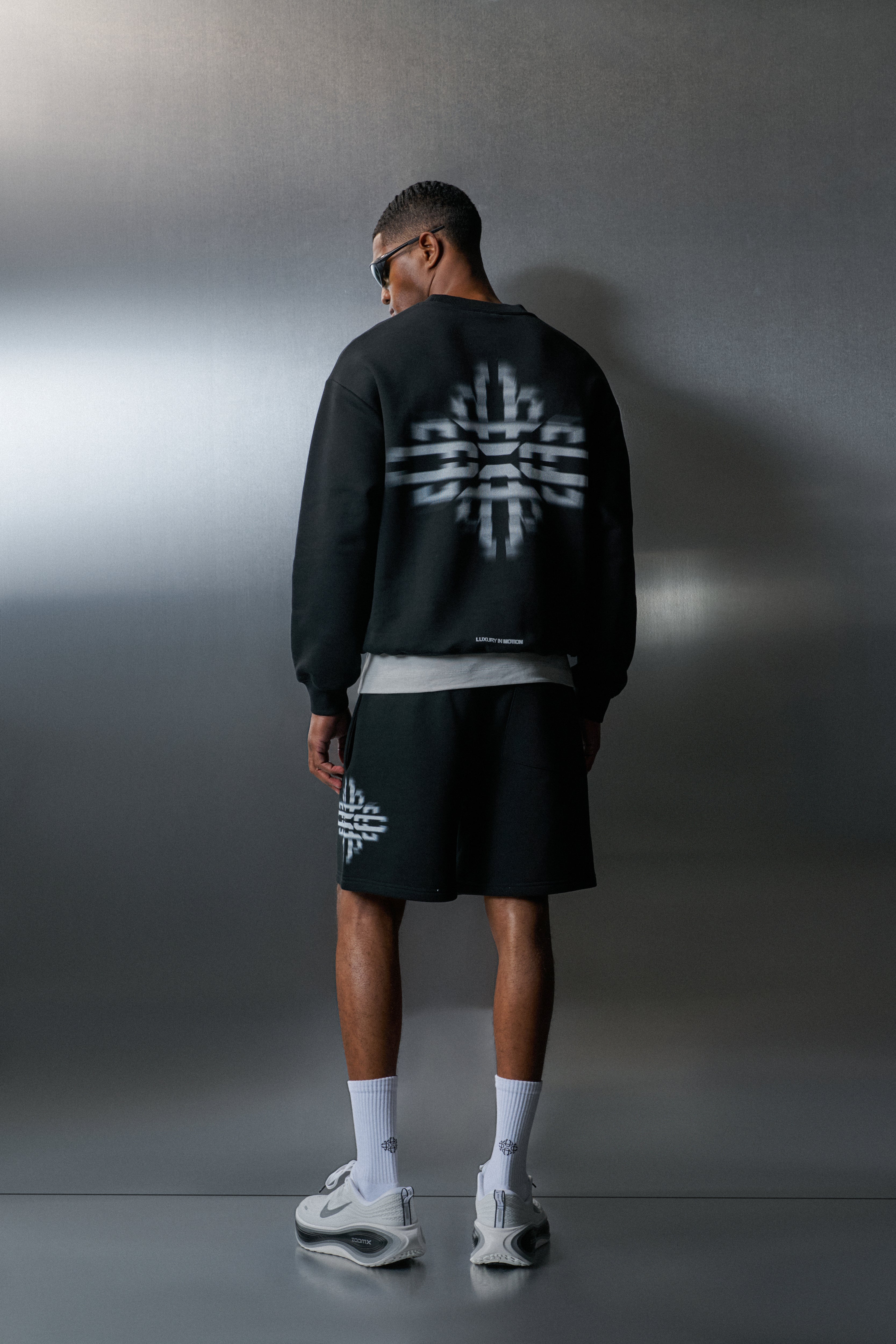 Person wearing a black sweatshirt and shorts with a logo, standing against a grey background