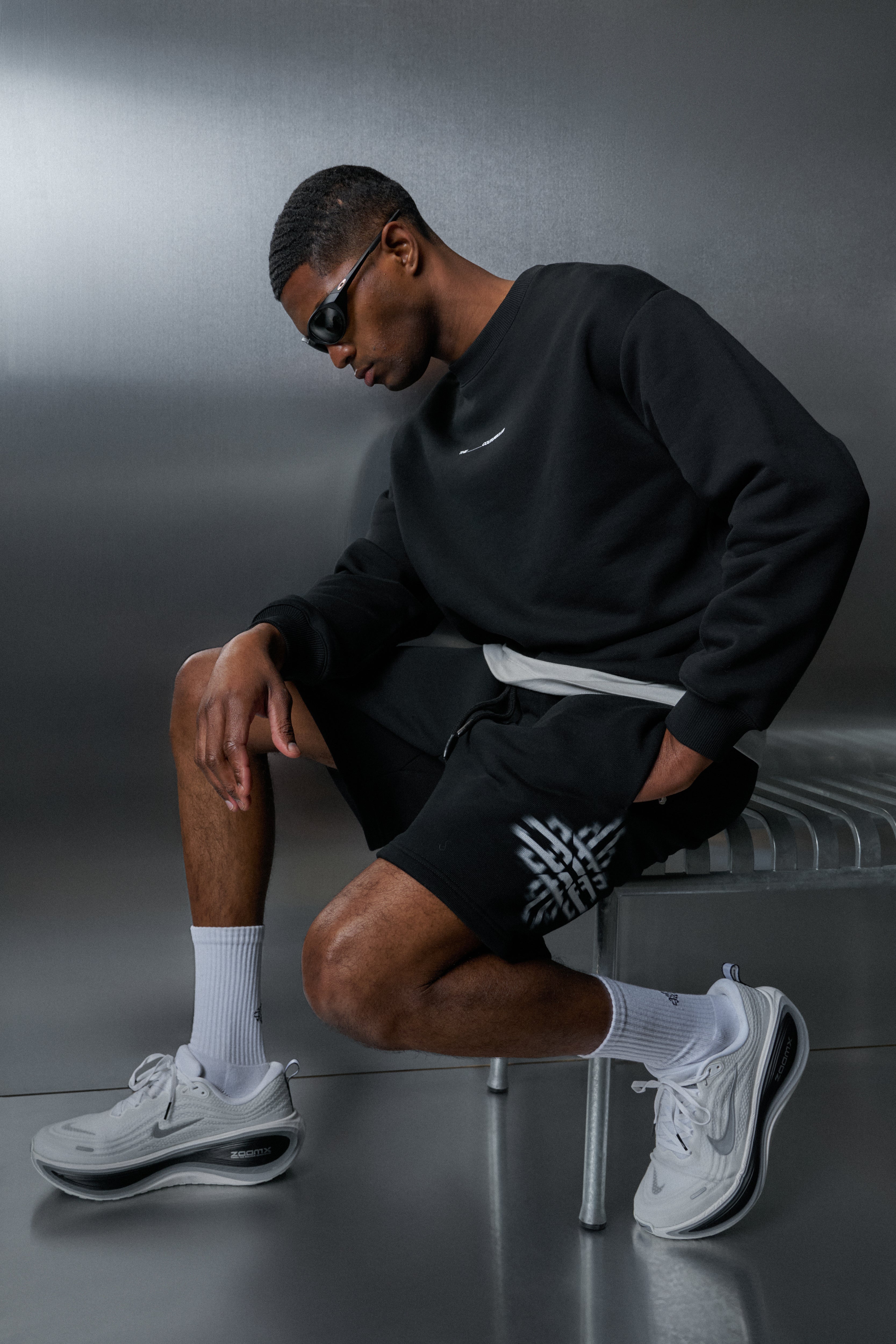 Man wearing black sweatshirt, black shorts with white logo, and white sneakers on a grey background