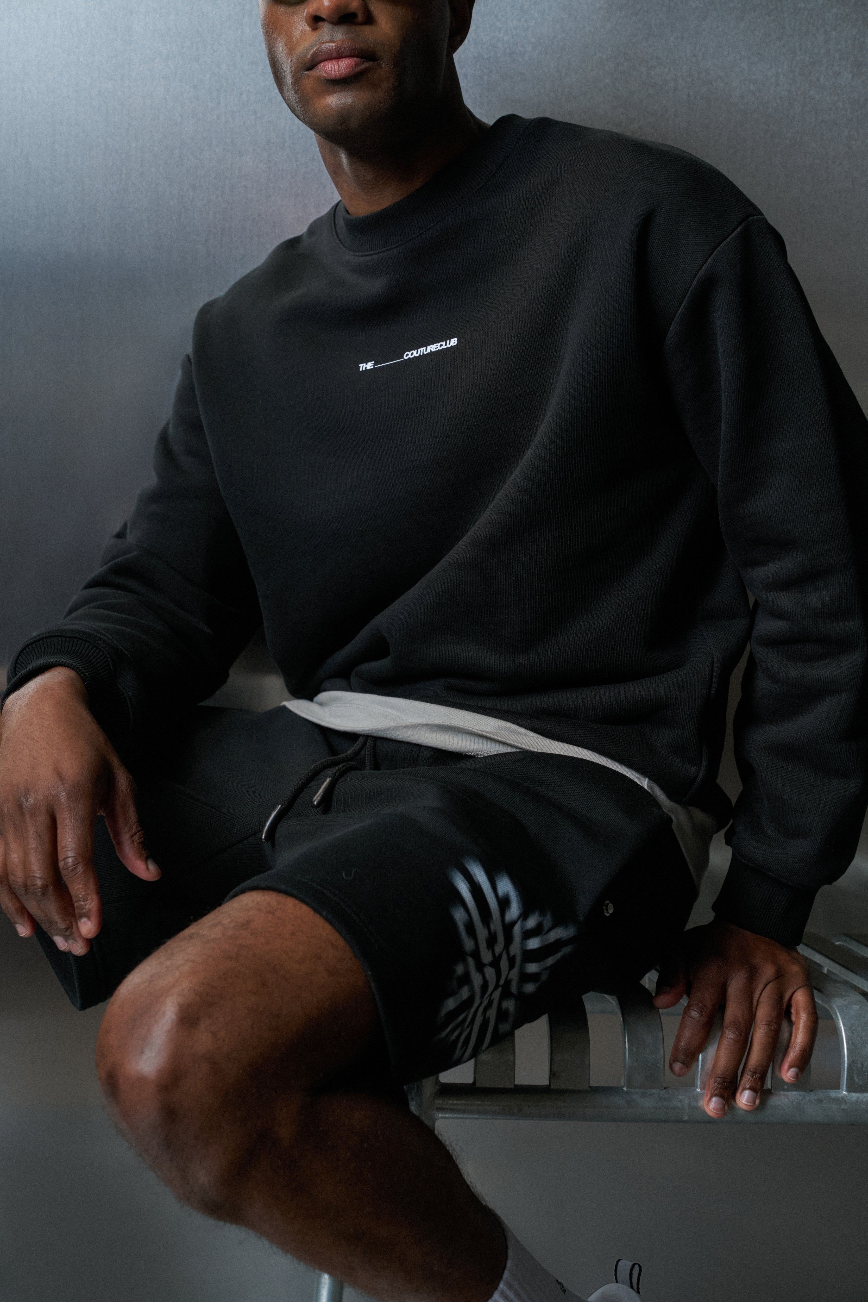 Person wearing a black sweatshirt and shorts sitting on a chair against a grey background