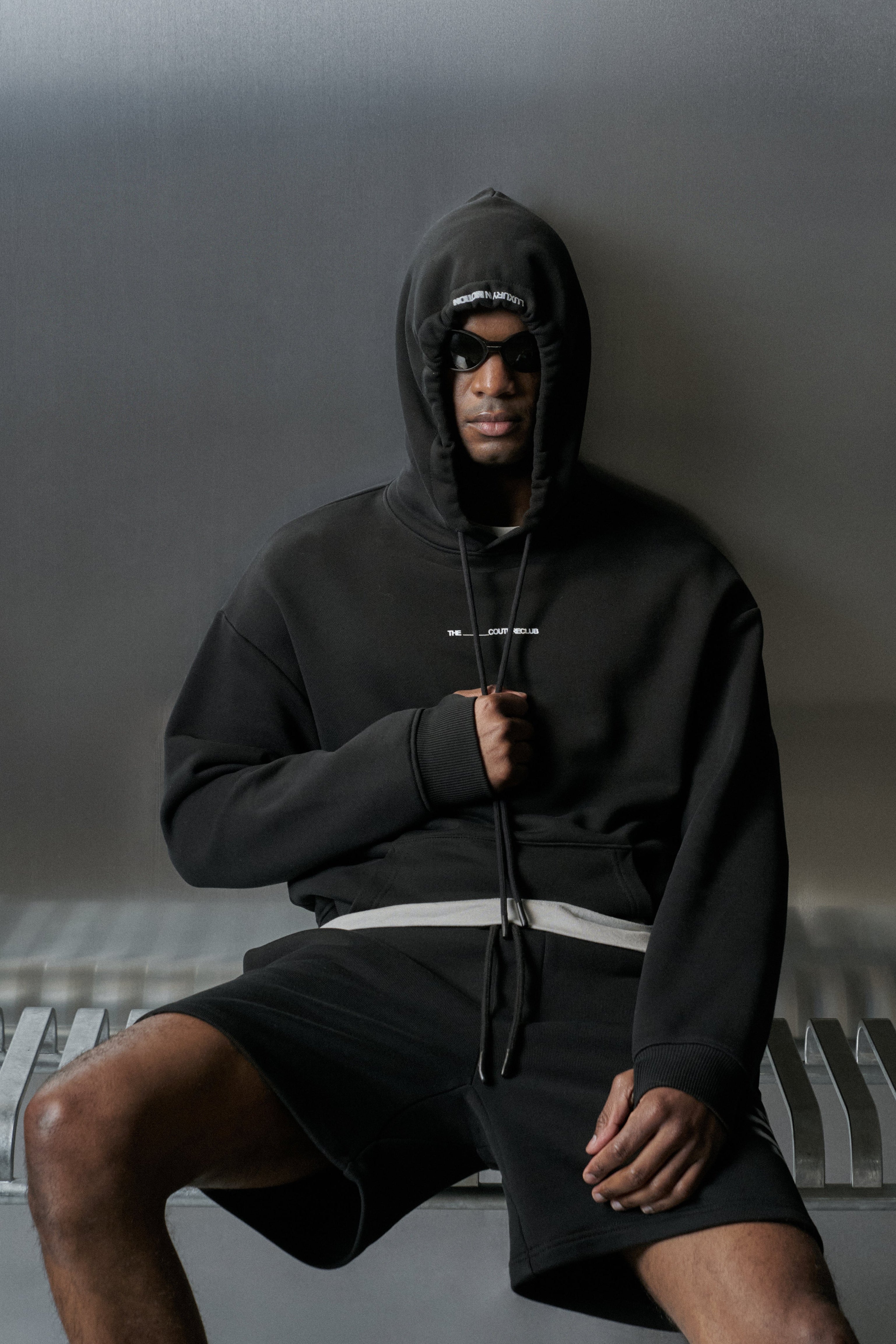Person wearing a black hoodie and shorts sitting on a bench against a grey background
