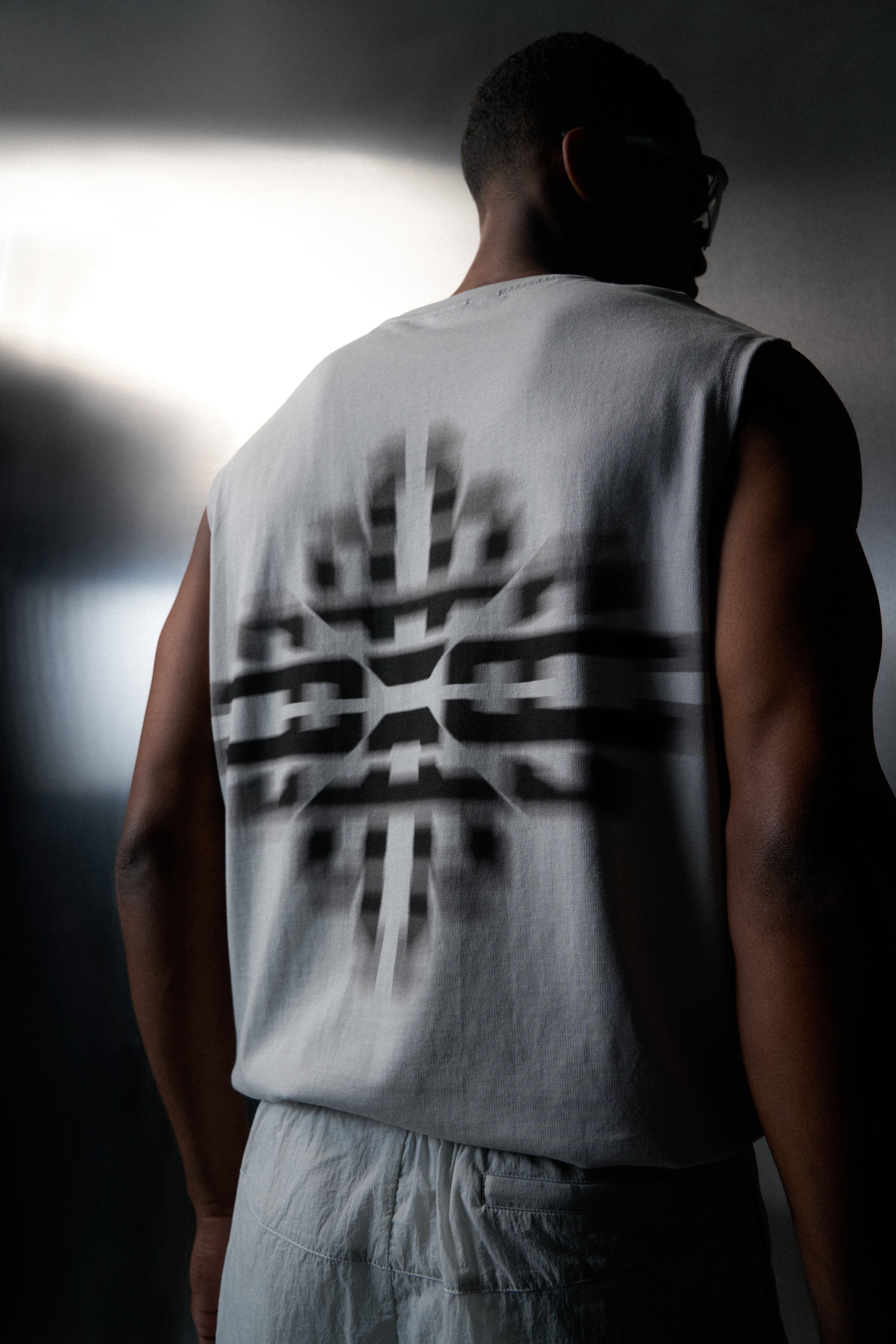 Person wearing a white sleeveless shirt with a logo on the back, standing against a dark background.