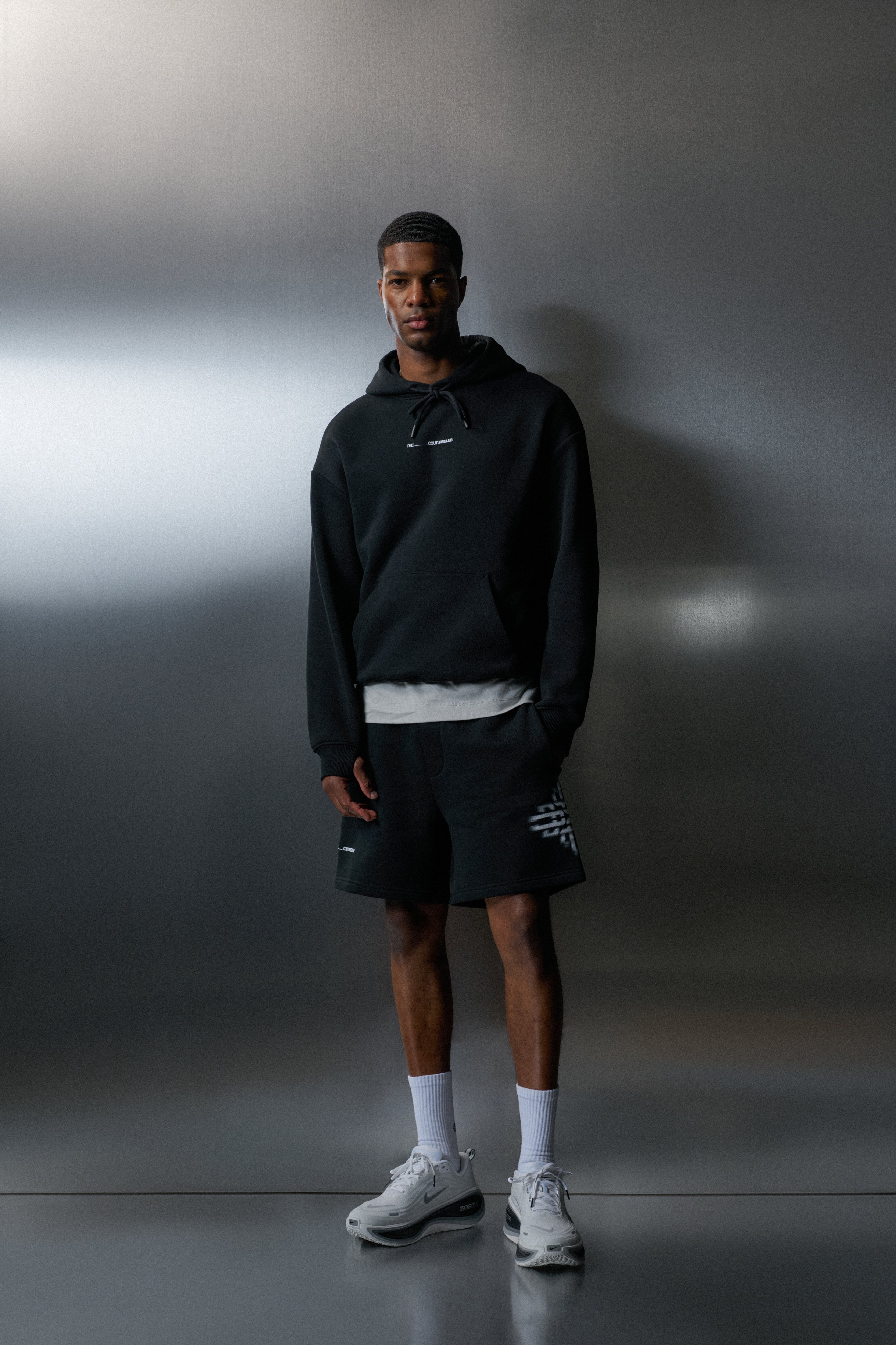 Man wearing a black hoodie and shorts with white sneakers against a grey background