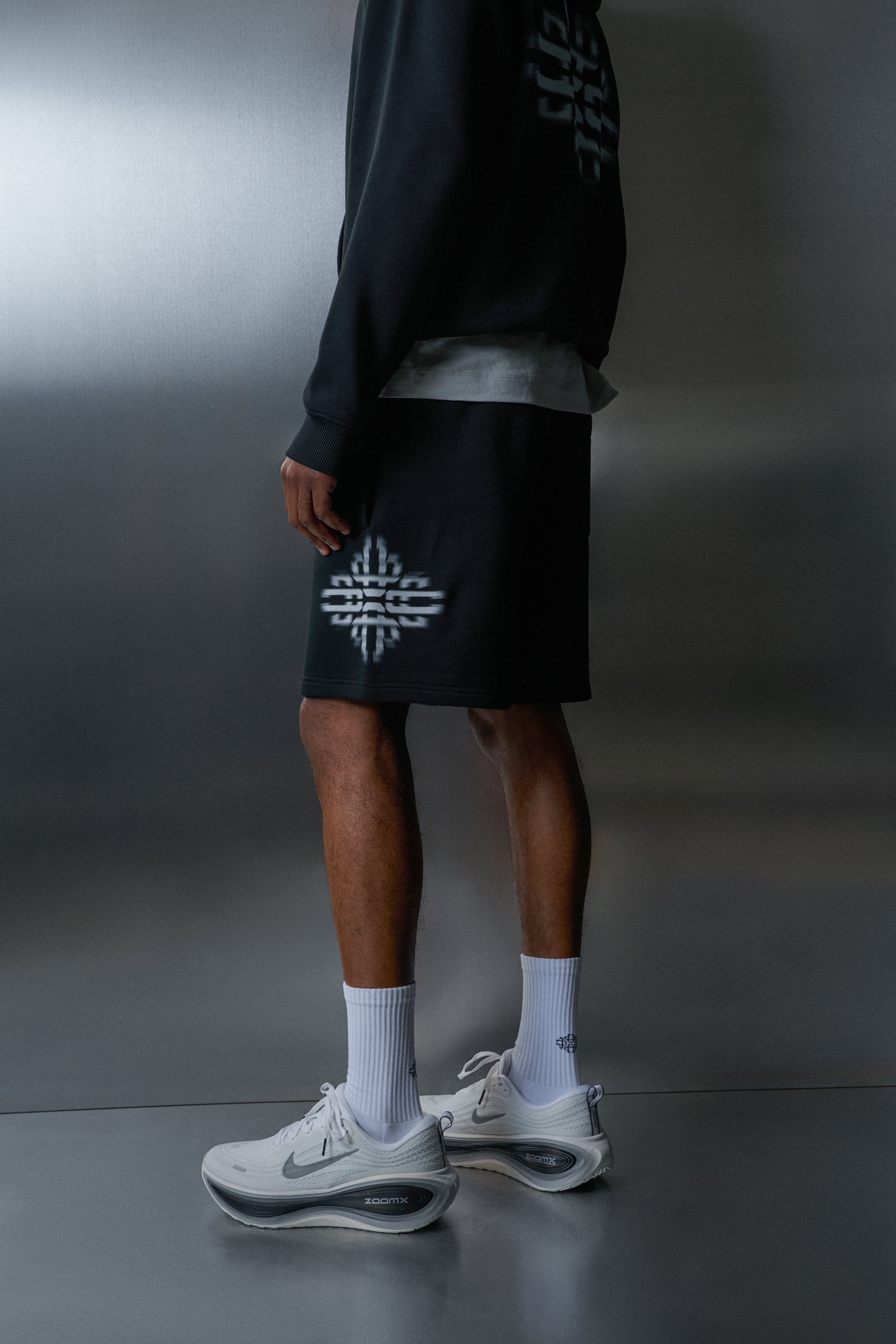 Person wearing black shorts with a logo, white socks, and grey sneakers on a dark background