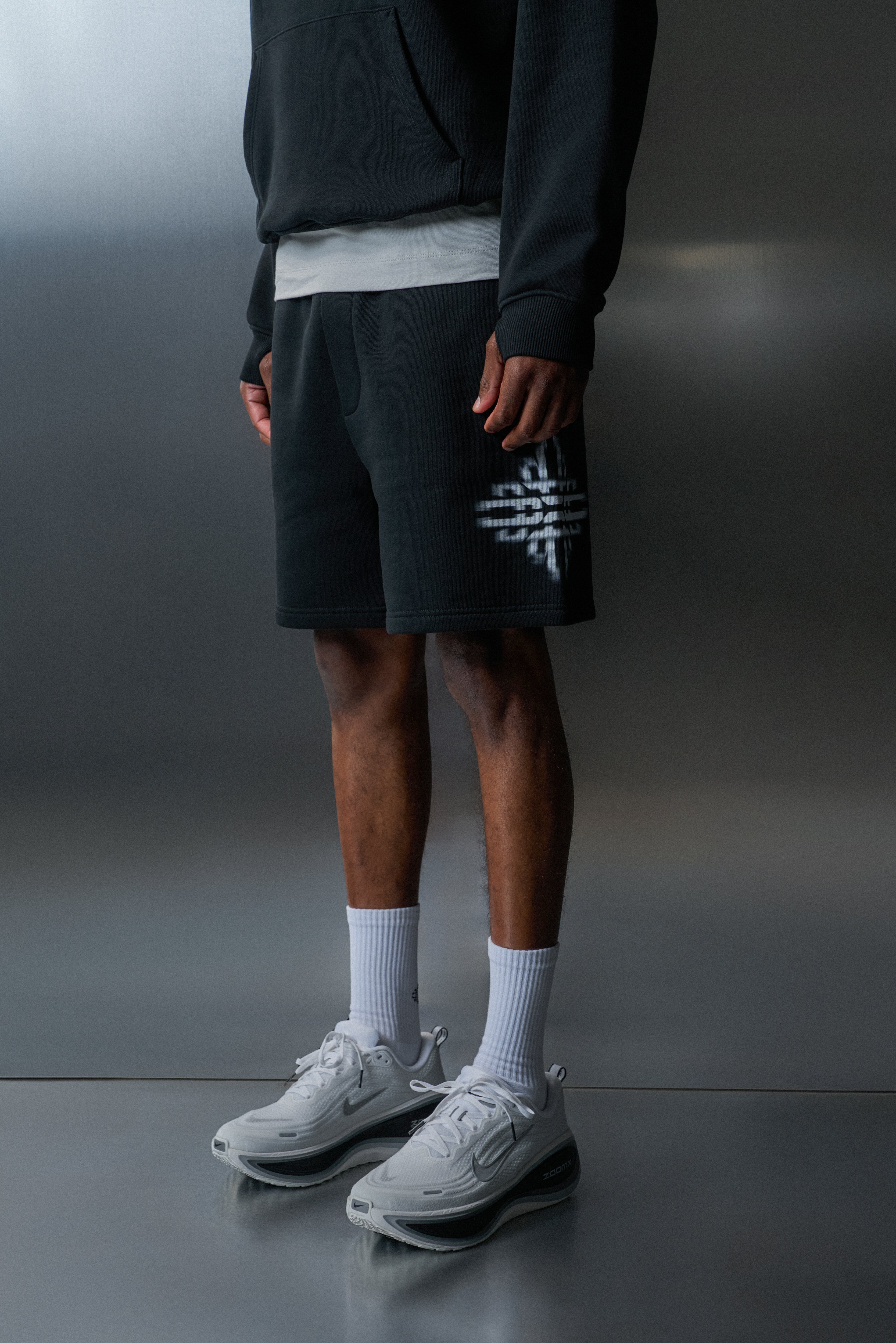 Person wearing black shorts with a logo, white sneakers, and socks on a grey background