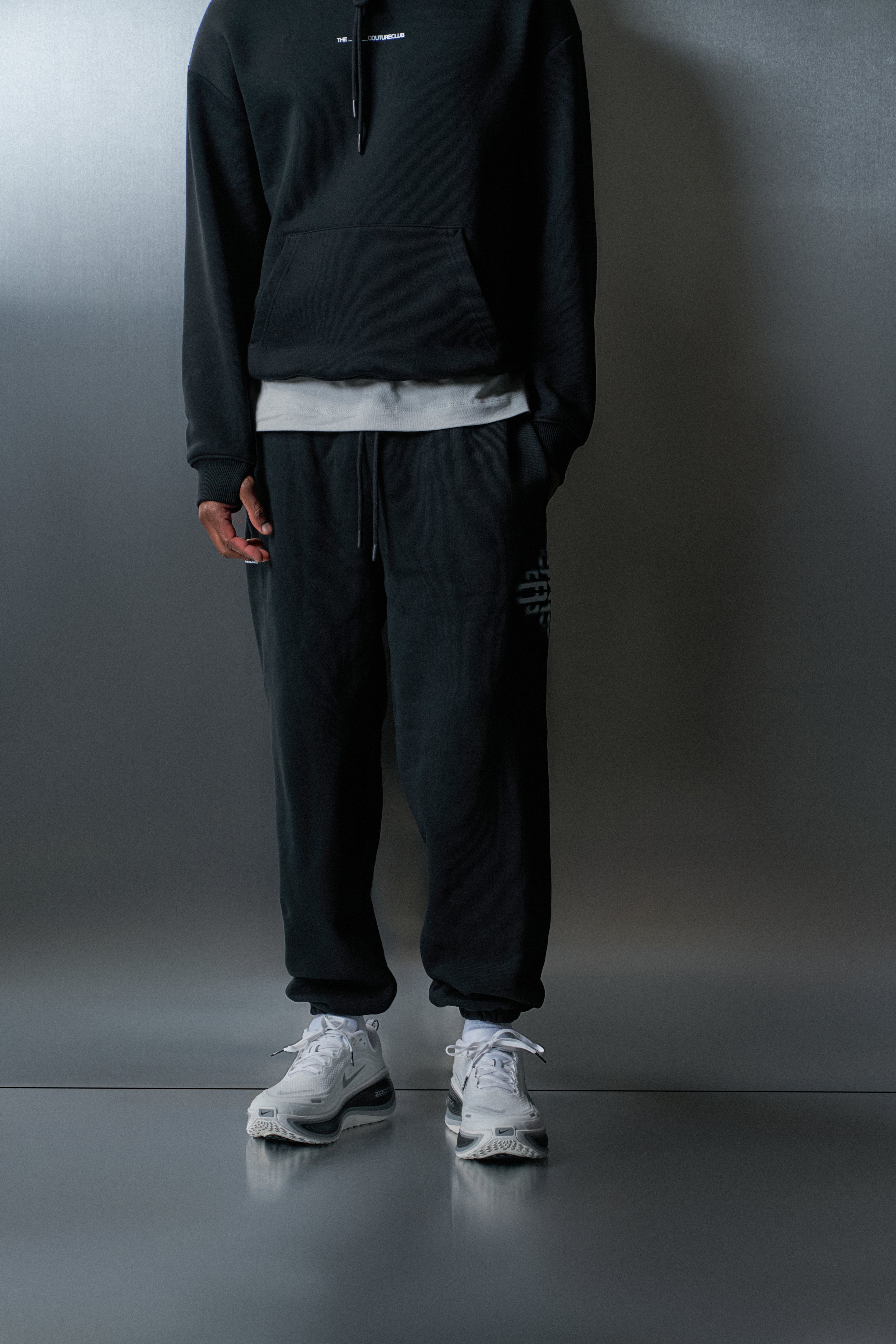 Person wearing a black hoodie and joggers with white sneakers against a grey background