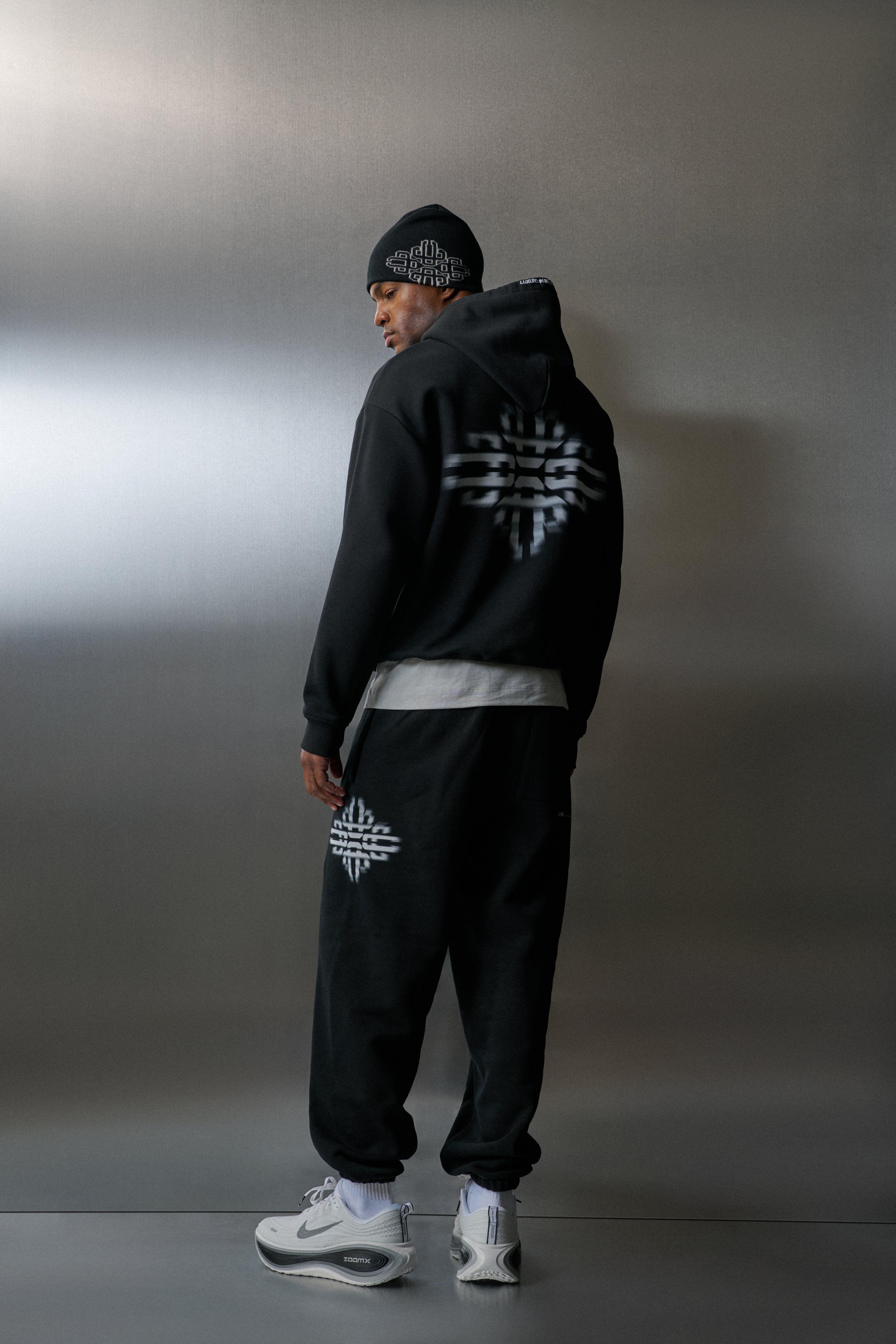 Person wearing a black hoodie and pants with a logo, standing against a grey background