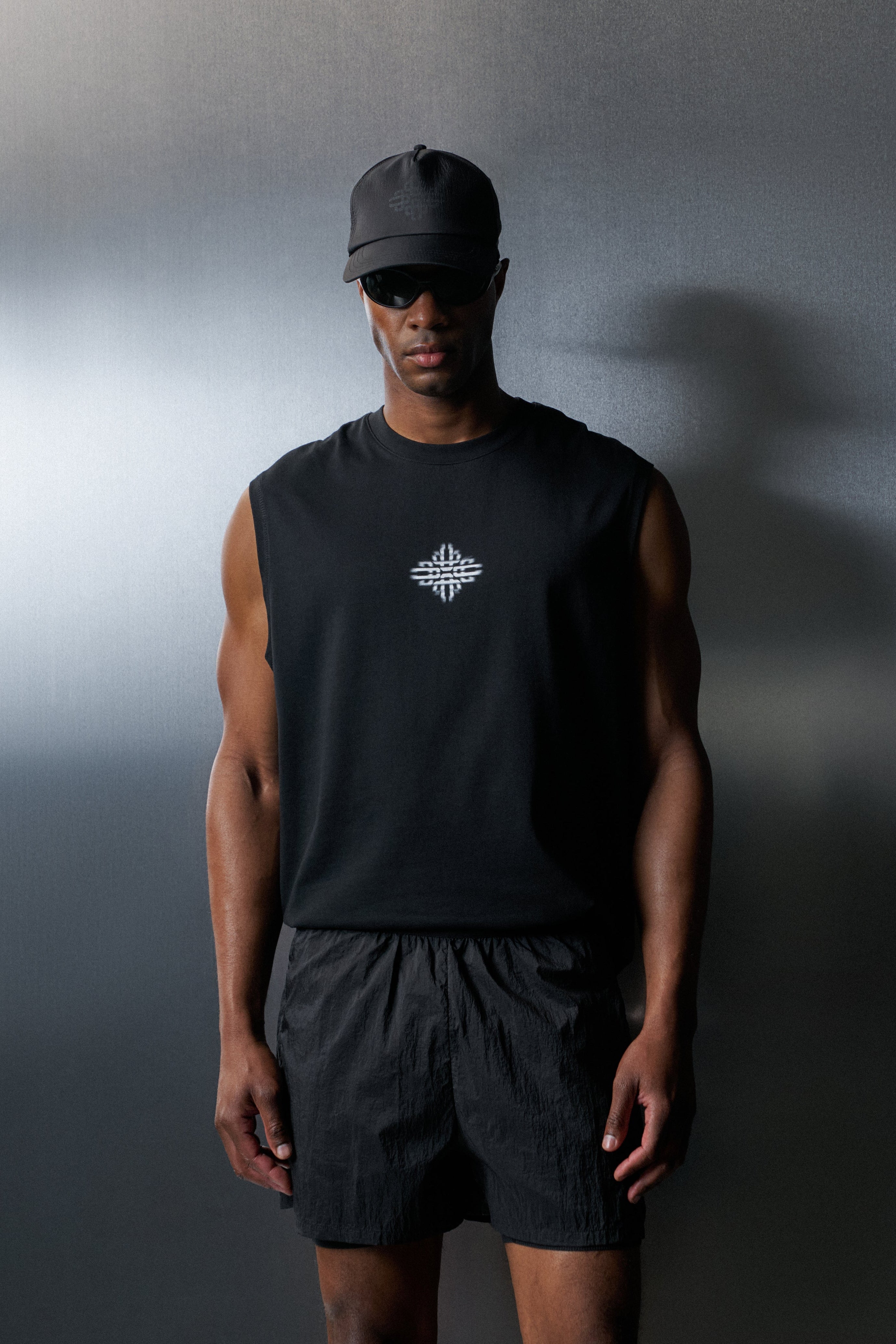 Man wearing a black sleeveless shirt and shorts with a logo, standing against a gradient grey background.
