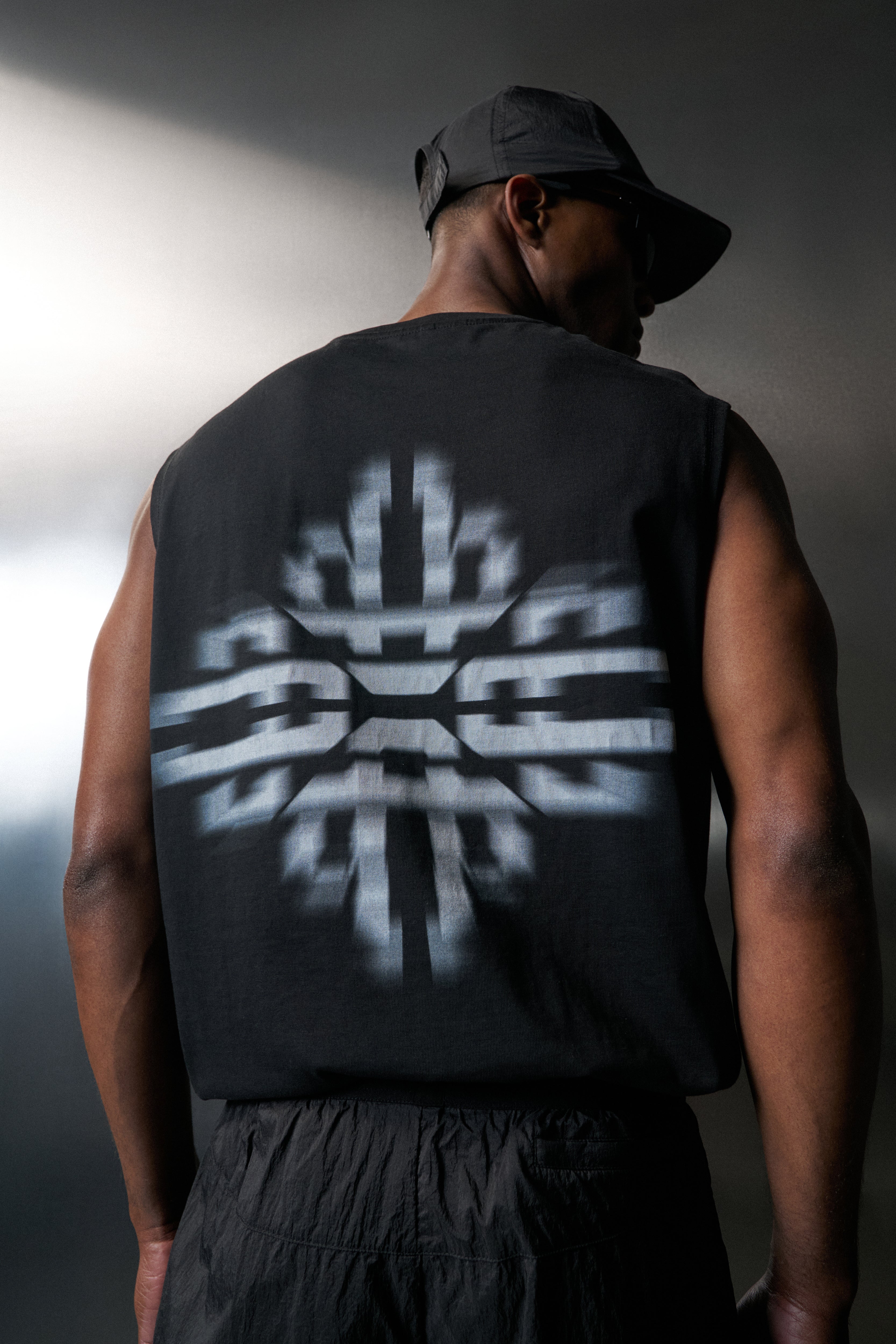Person wearing a black sleeveless shirt with a geometric design on the back, standing against a dark background.