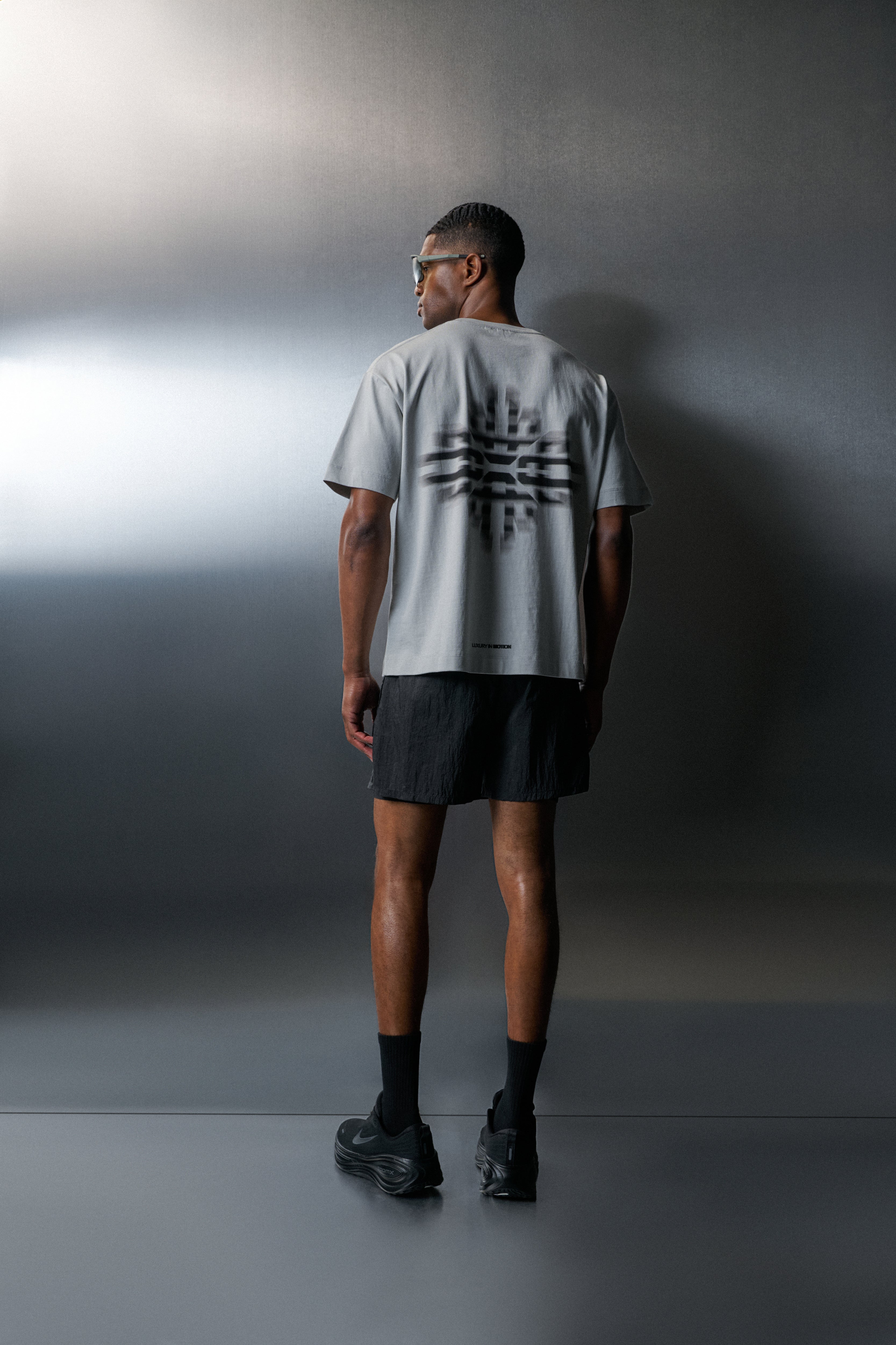 Man wearing a light grey t-shirt with a black design on the back, standing against a grey wall.