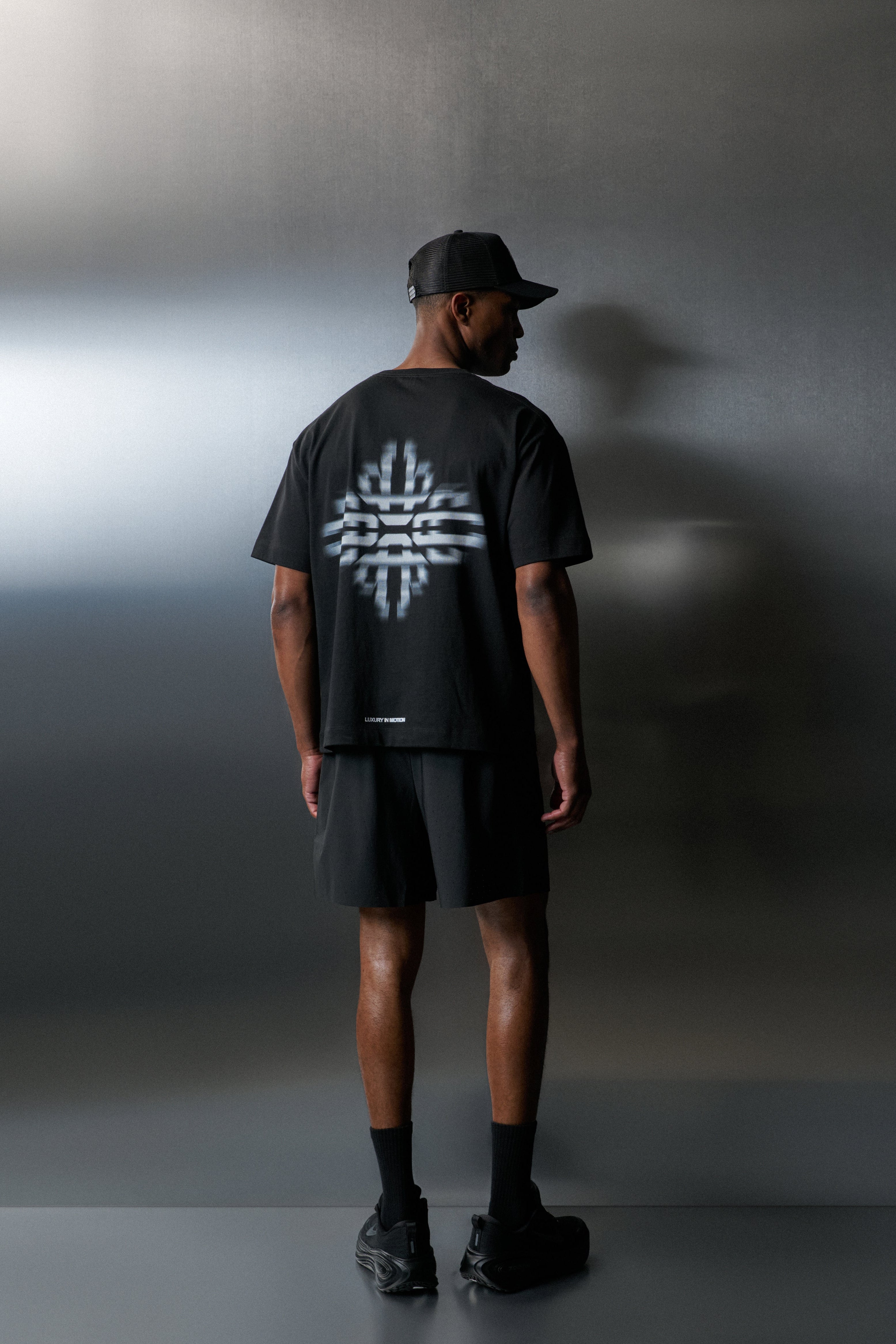 Person wearing a black t-shirt with a white logo on the back, standing against a grey wall.