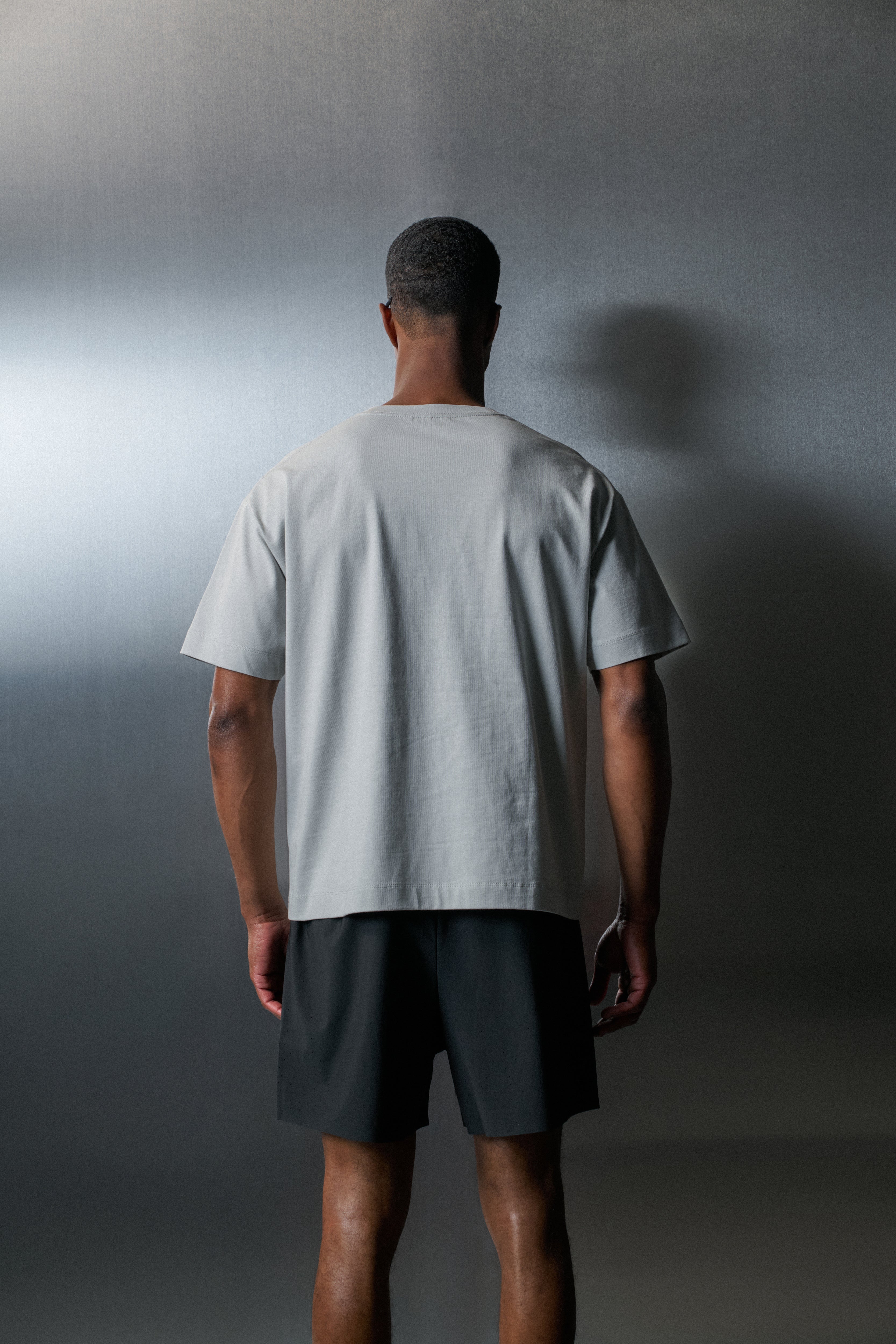 Person wearing a light grey t-shirt and dark shorts standing against a gradient grey background