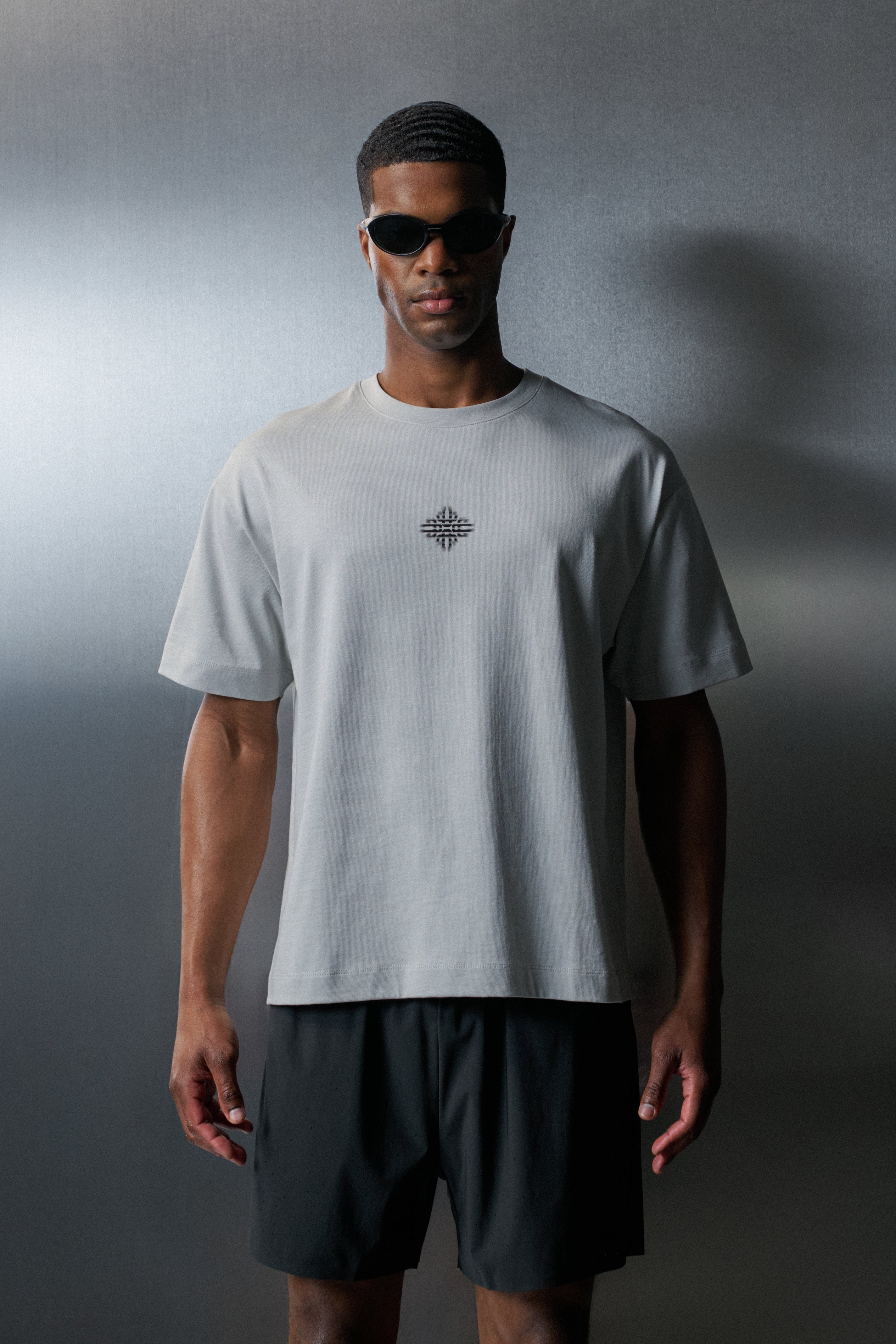 Man wearing a light grey t-shirt with a logo and dark shorts against a gradient grey background