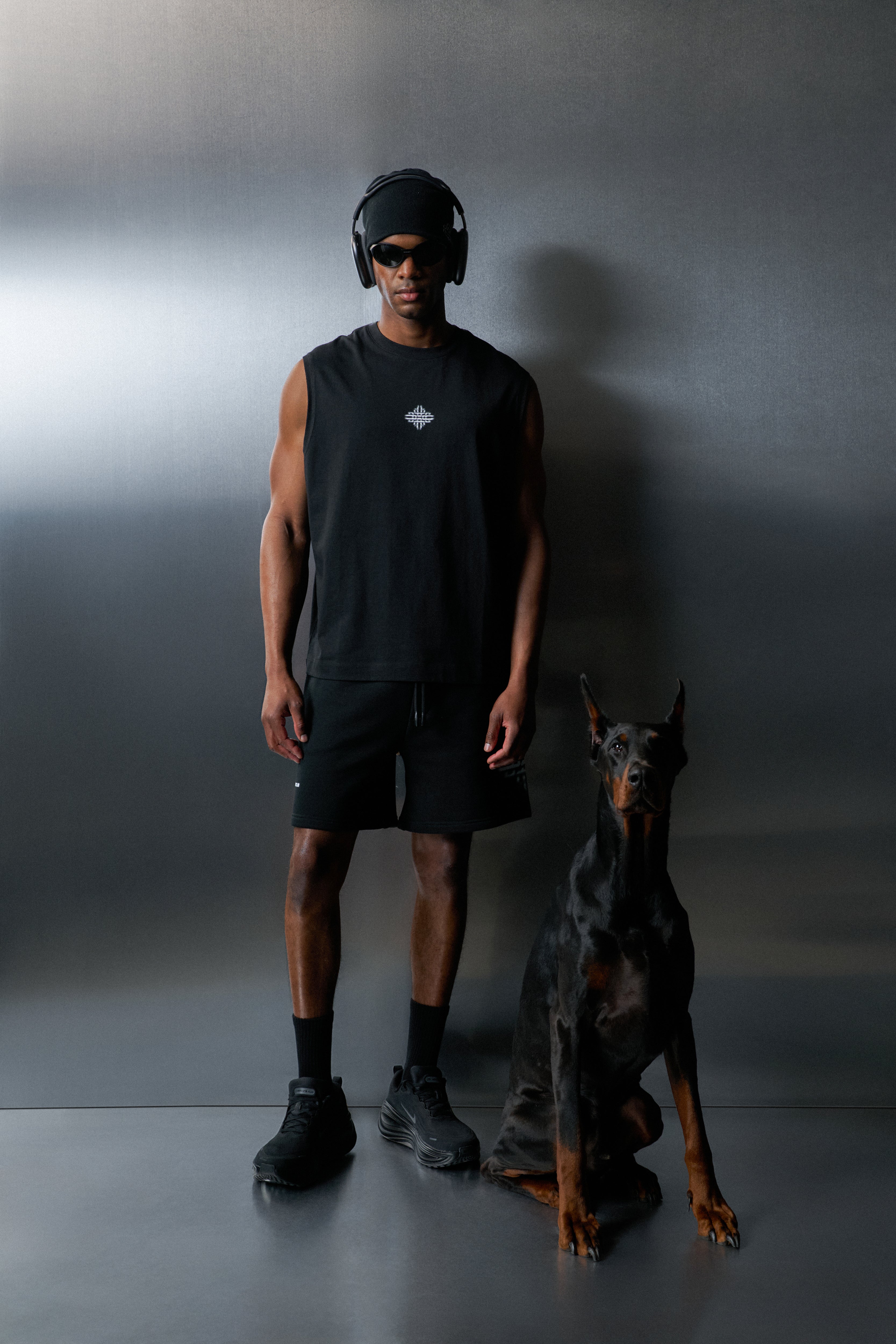 Man in black vest and shorts standing next to a Doberman dog against a grey background