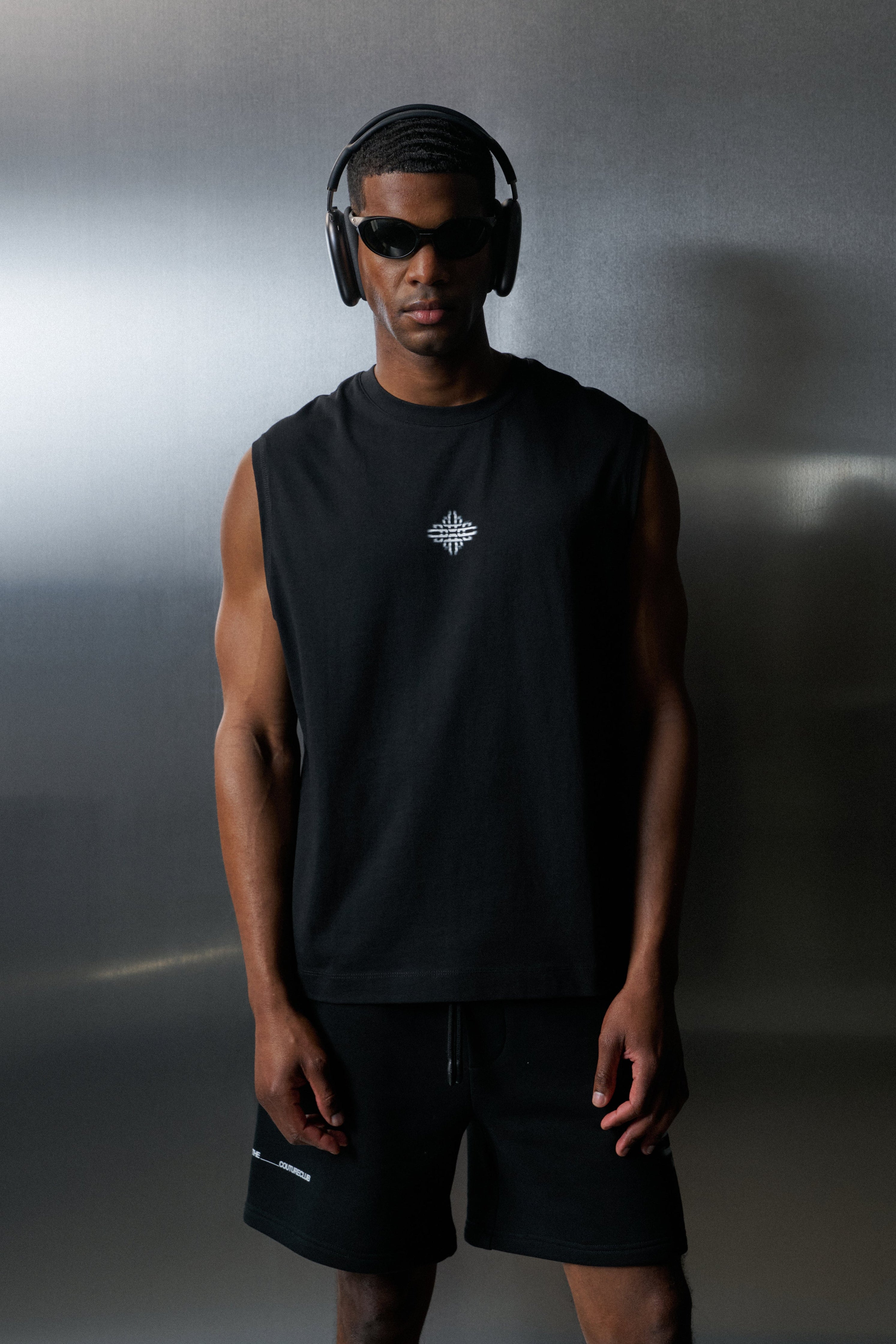 Man wearing black vest and shorts with headphones and sunglasses against a grey background