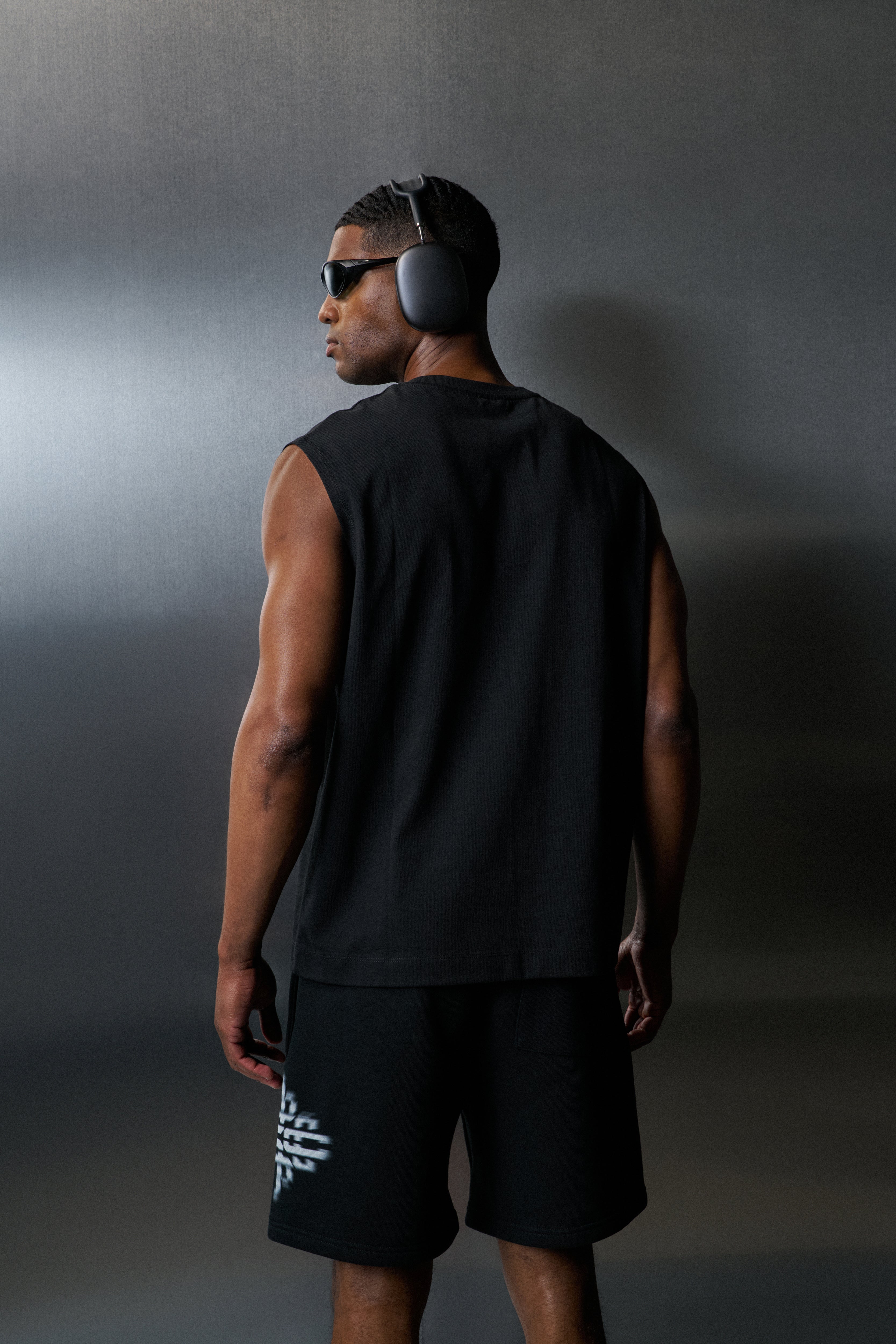 Man wearing black vest and shorts with sunglasses and headphones against a dark background