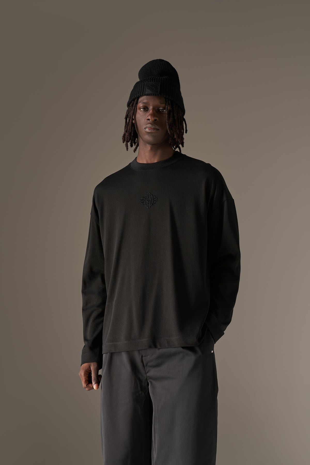 Man in a long sleeved black t-shirt, beanie and black pants against a neutral background
