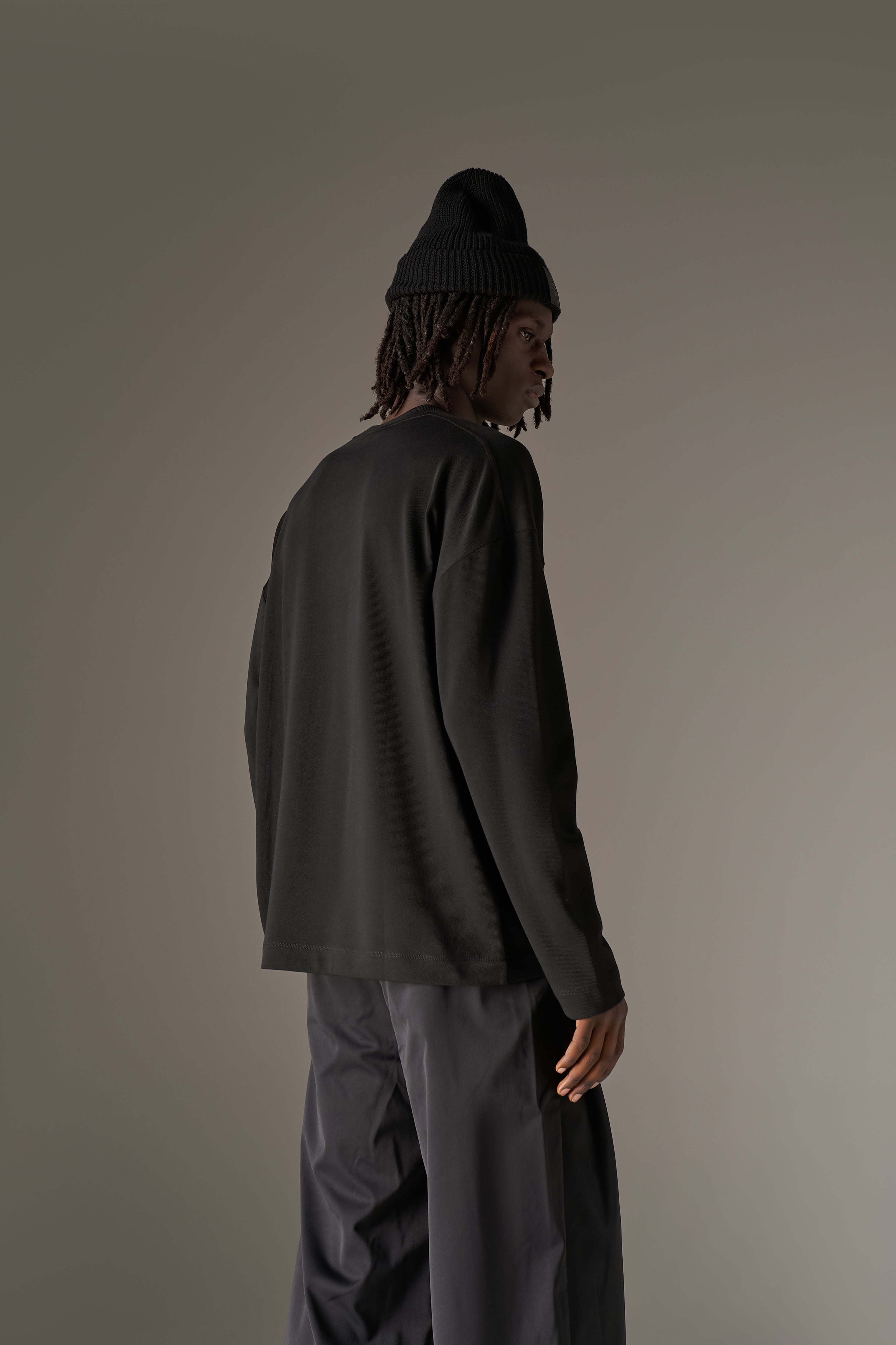 Back view of a man in a long sleeved black t-shirt, beanie and black pants against a neutral background.