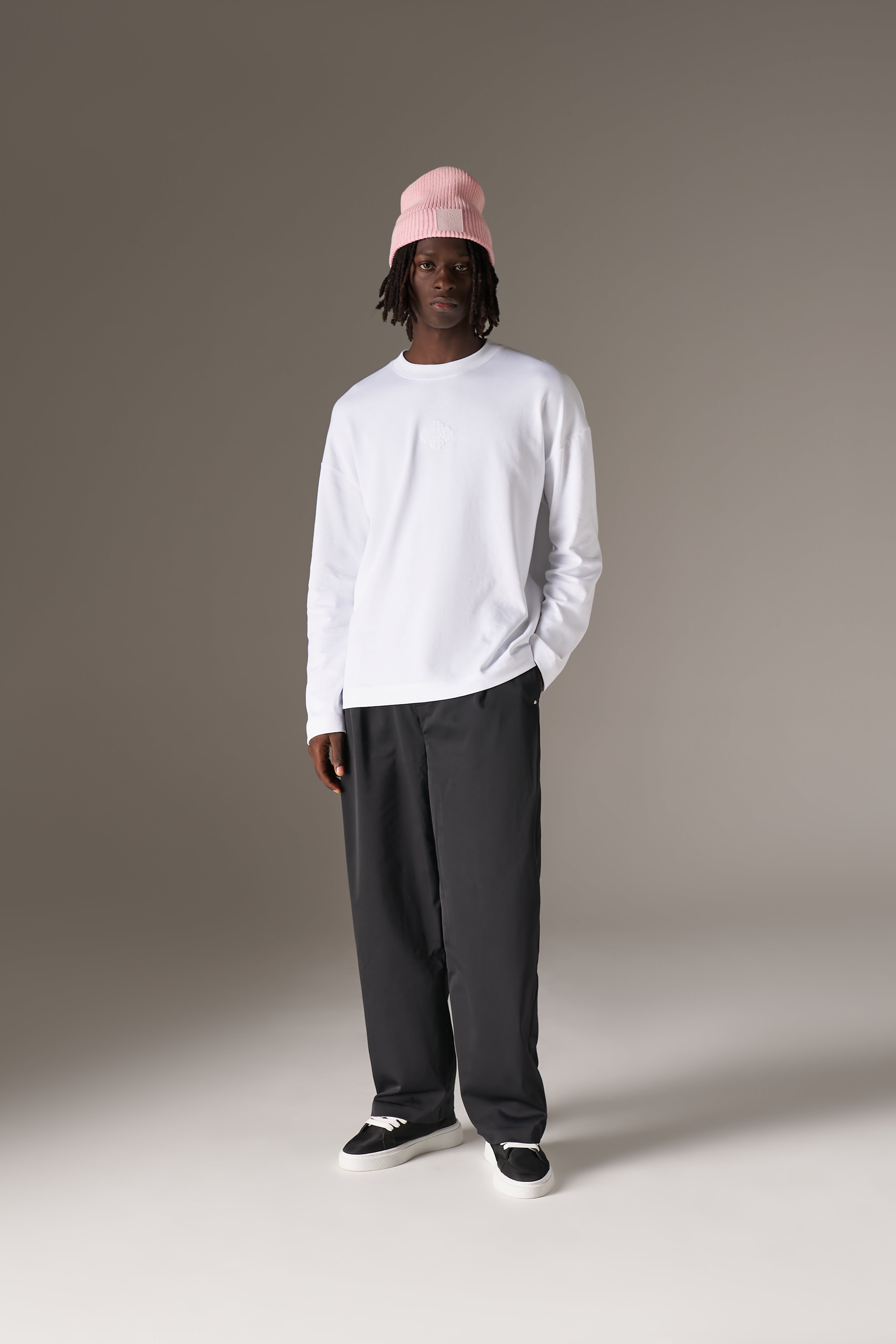 Person wearing a white long sleeve t-shirt, grey pants, and a pink beanie on a plain background