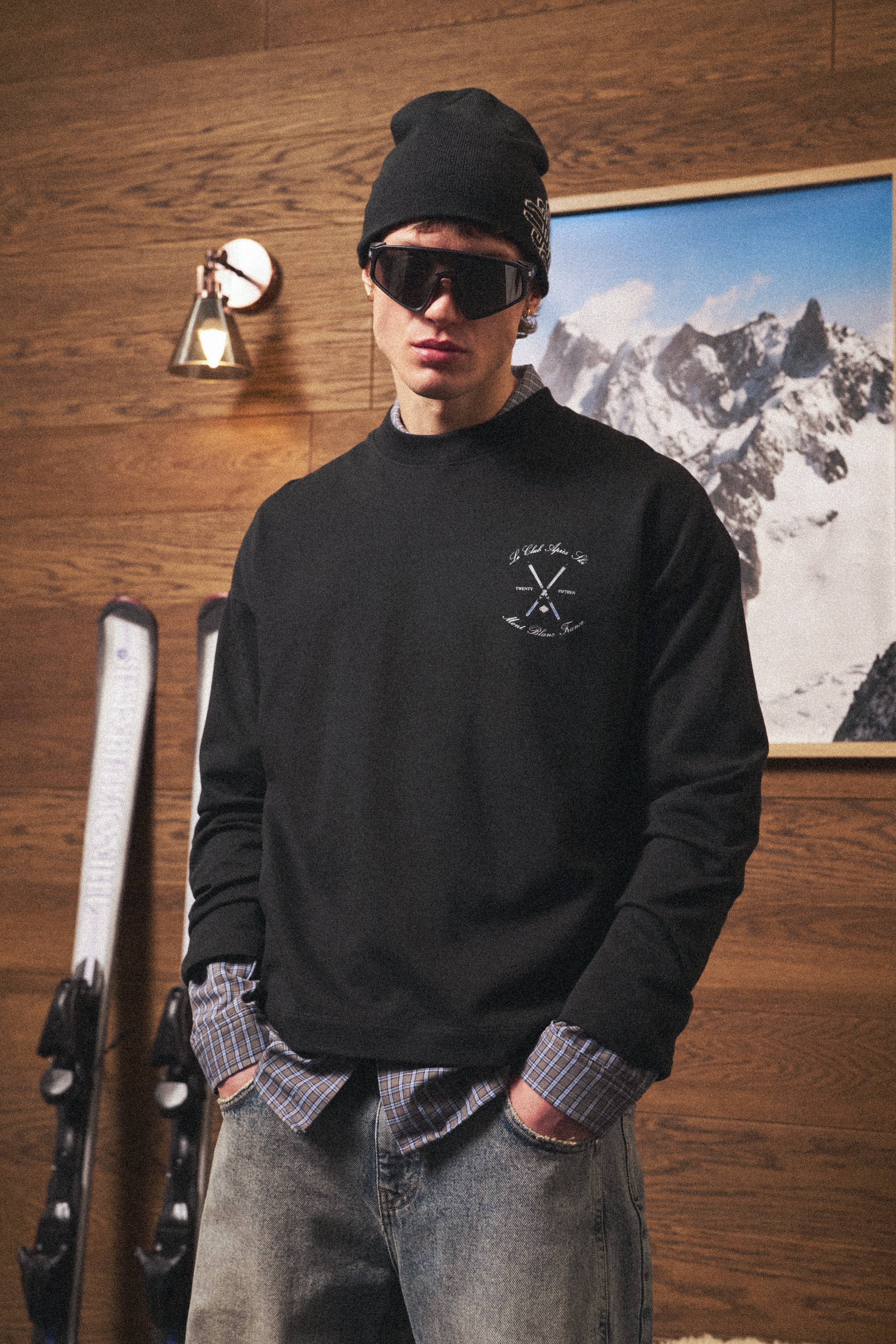 Person wearing a black long sleeved t-shirt with a logo, sunglasses, and a beanie in front of a wooden wall with skis and a mountain picture.