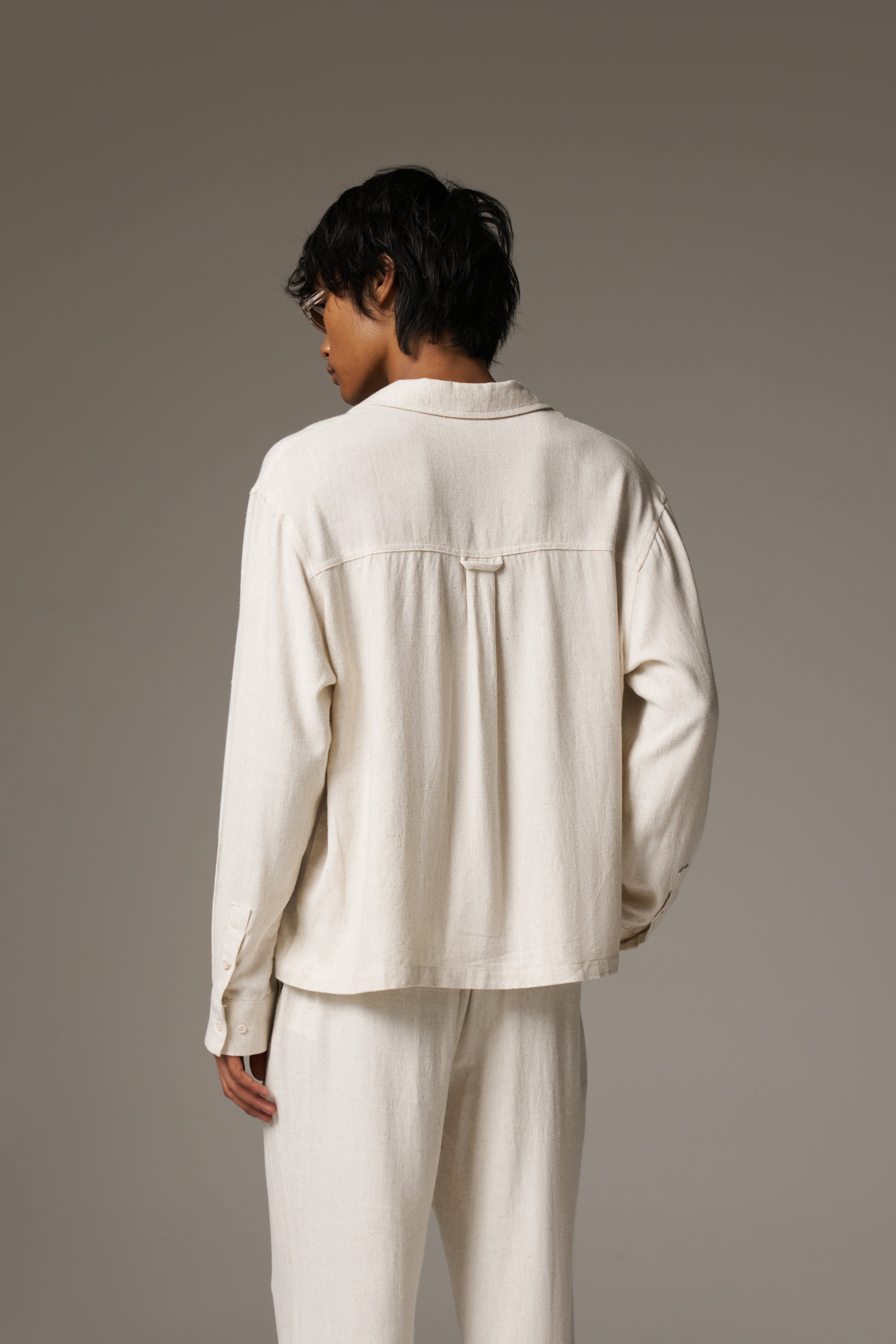 Back image of a man wearing a long sleeved linen shirt and patching pants, against a neutral background.