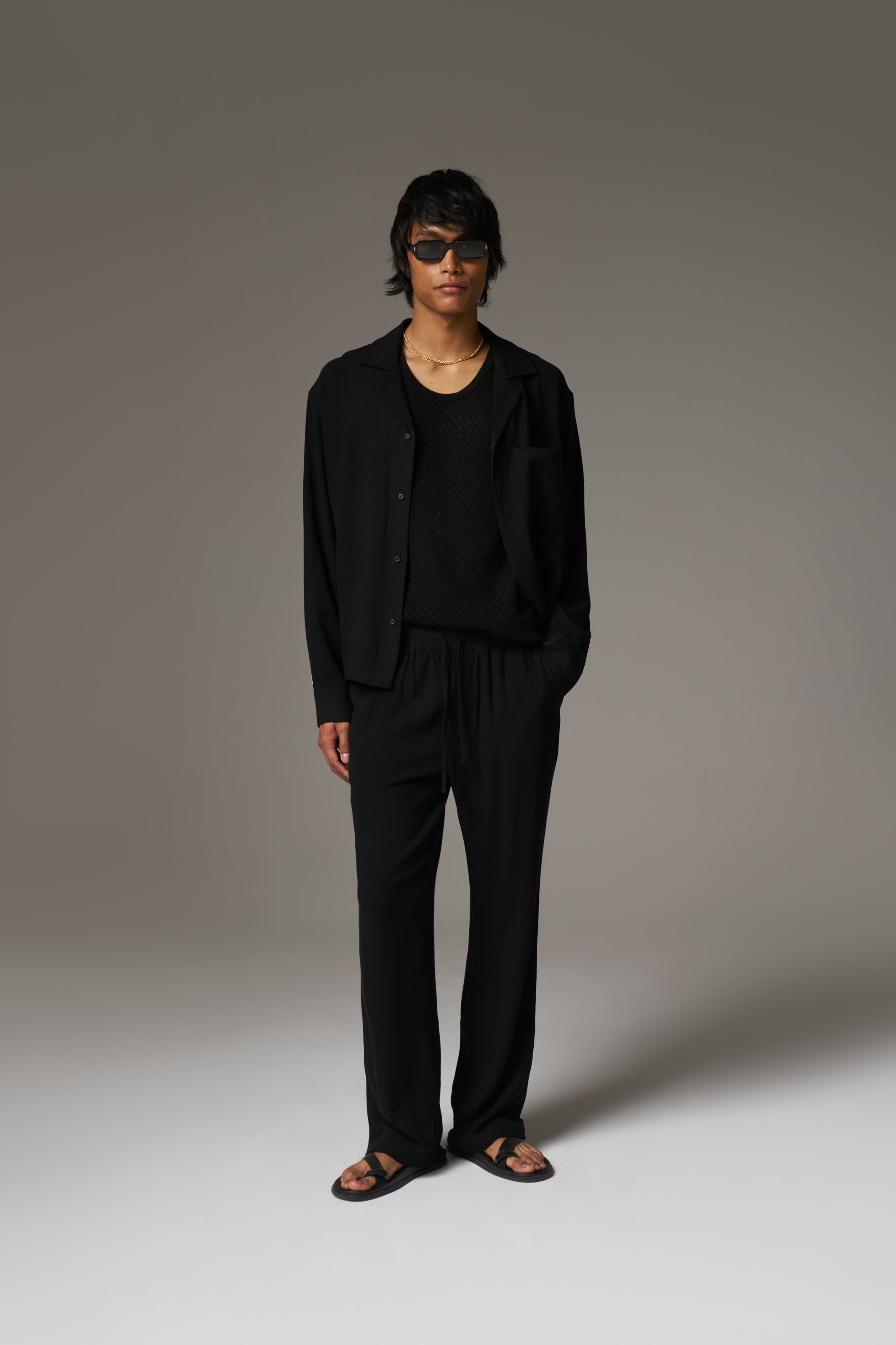 A man wearing a long sleeved shirt, with black sunglasses and pants against a neutral background.