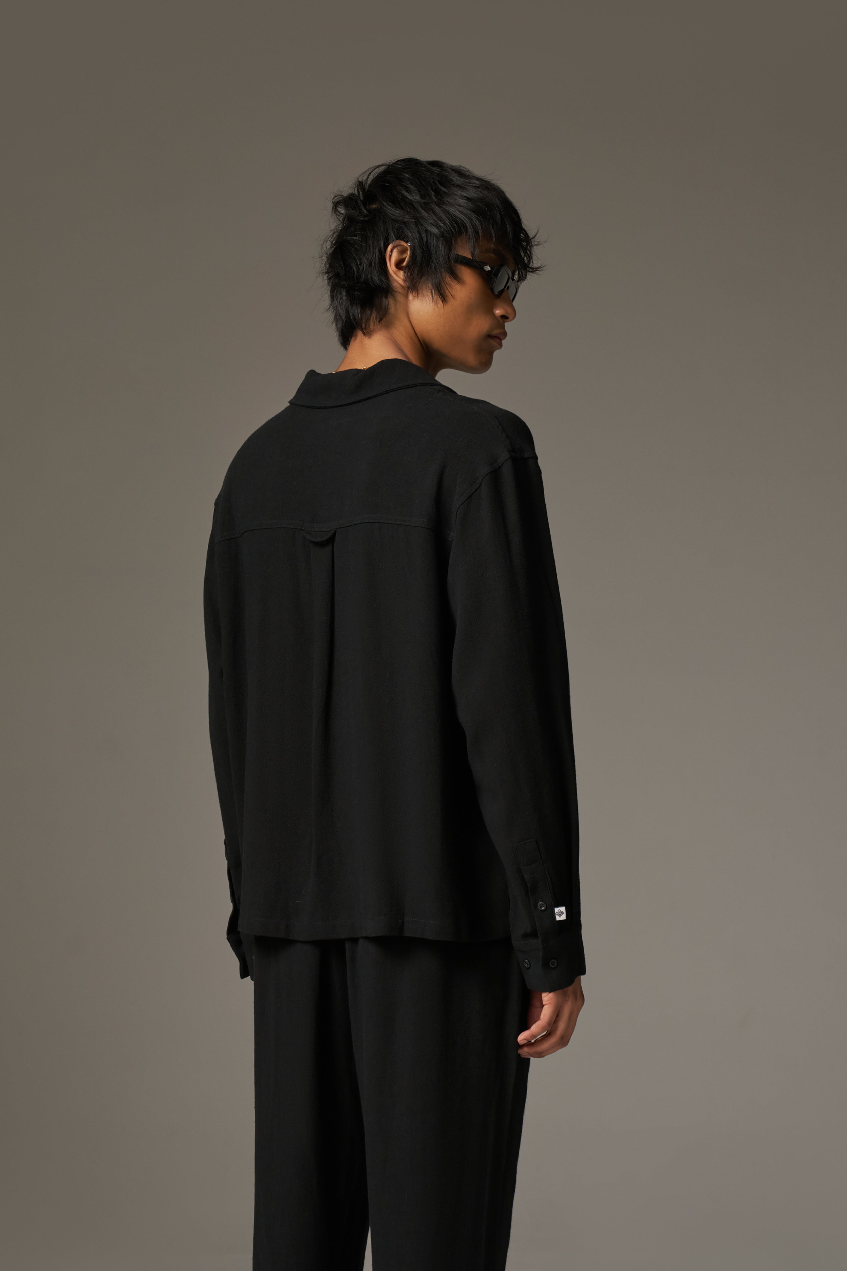 The back image of a man wearing a long sleeved shirt, with black sunglasses and pants against a neutral background.
