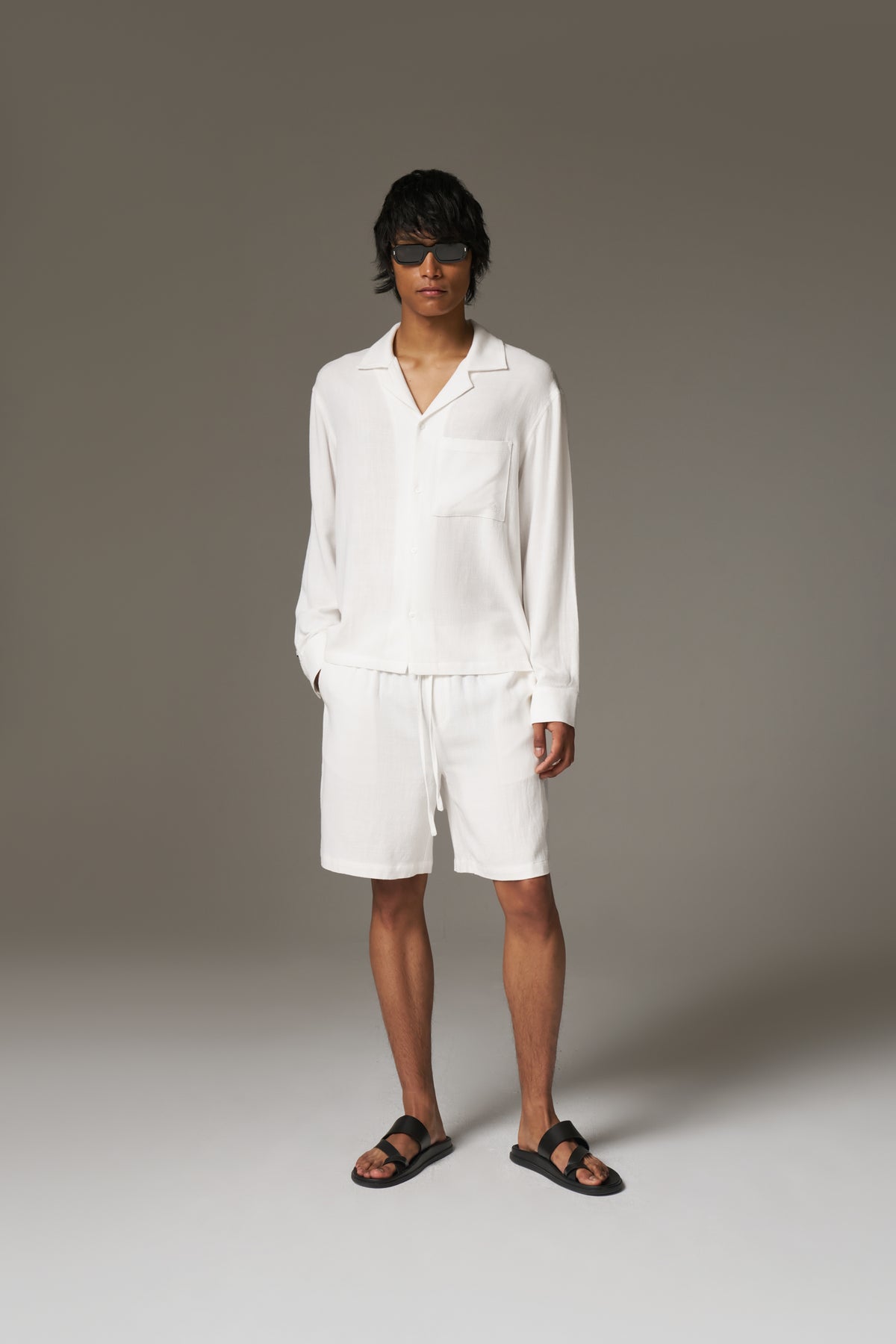 Man wearing a long sleeved white shirt, shorts and sunglasses against a neutral background.