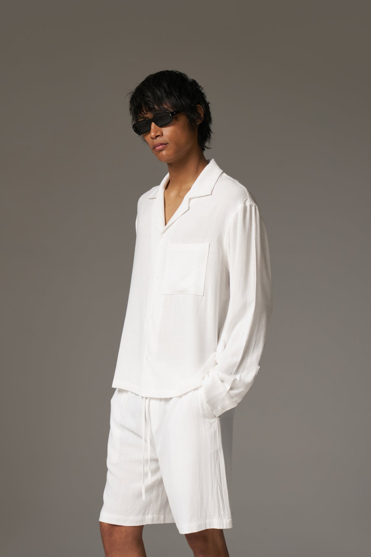 Man wearing a long sleeved white shirt, shorts and sunglasses against a neutral background.