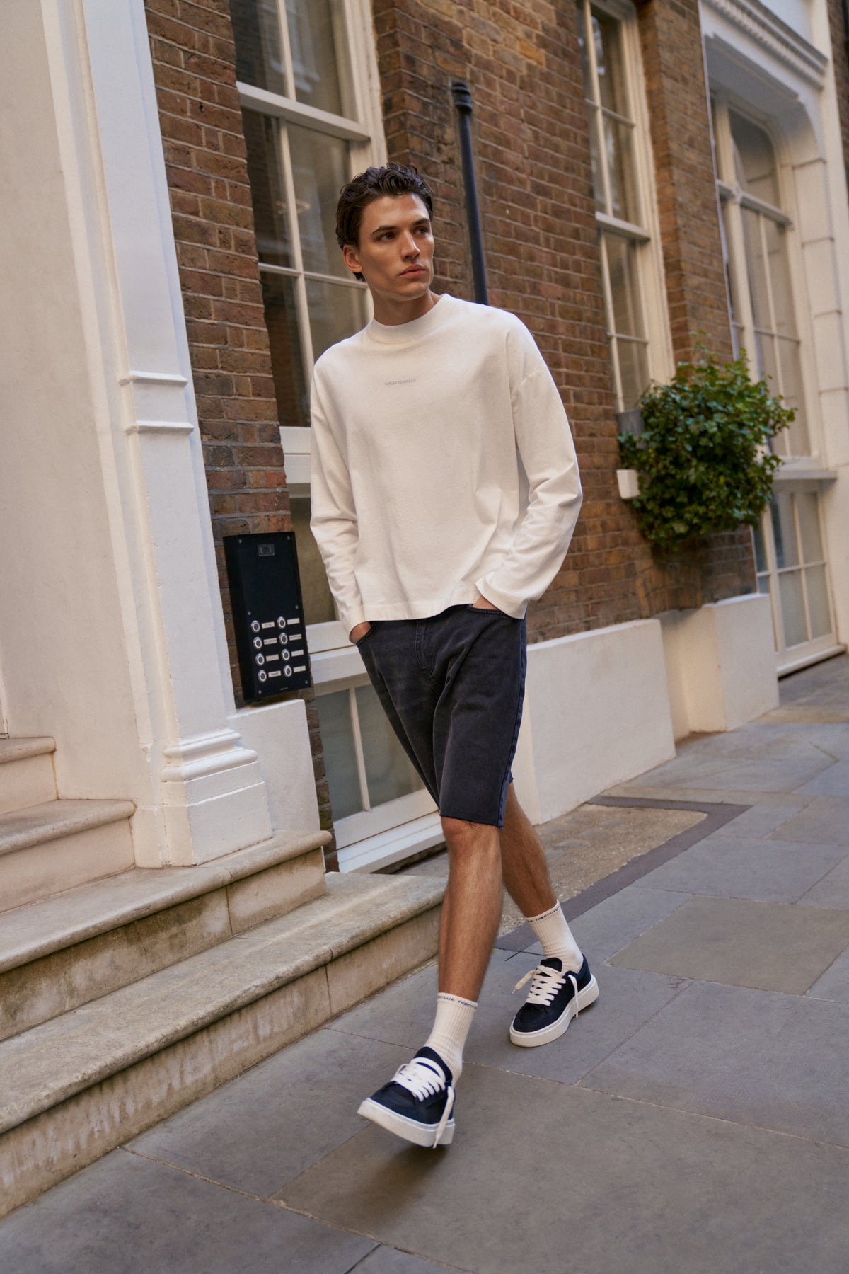 Man wearing a white t-shirt and black shorts standing on a sidewalk in an urban setting.
