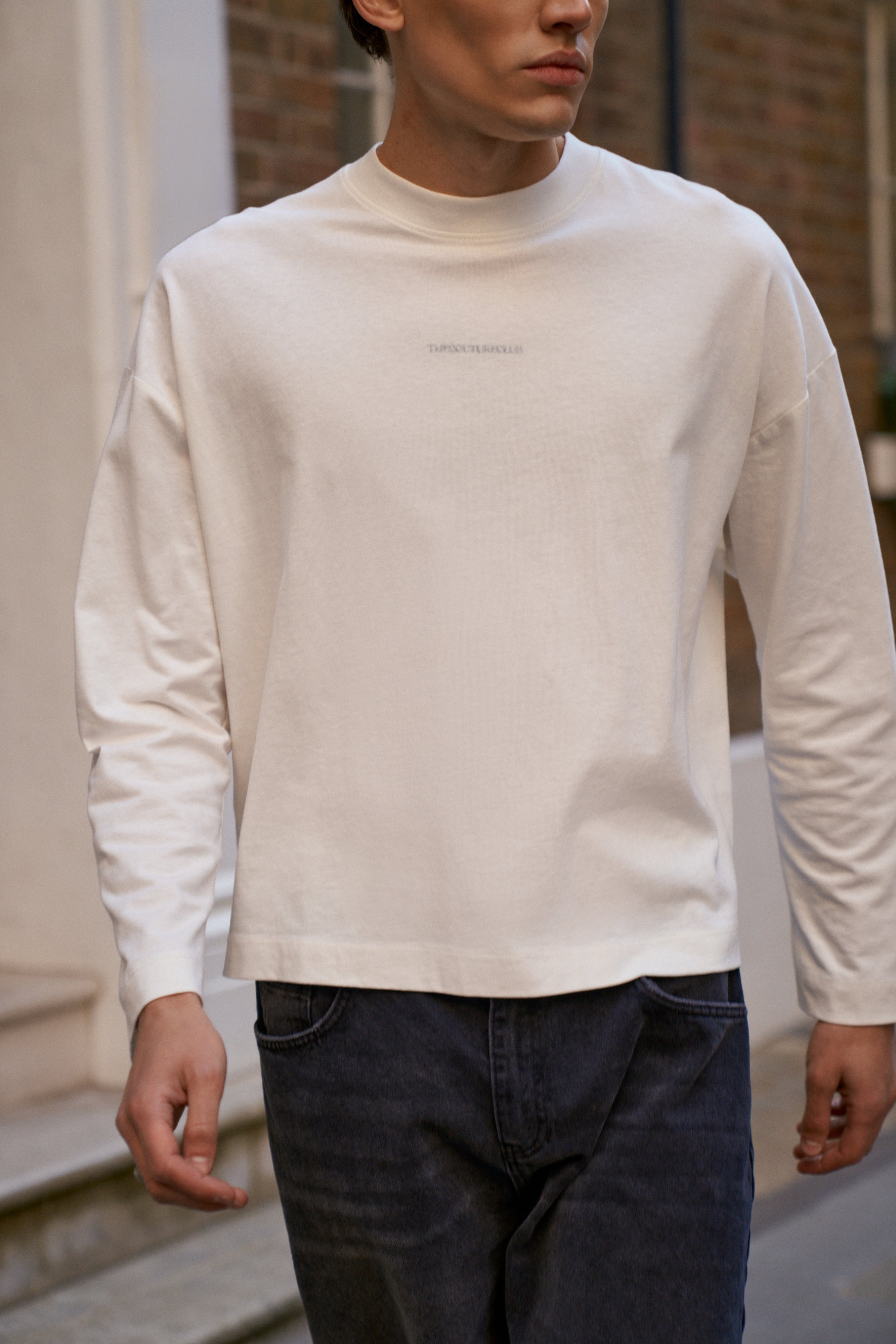 Person wearing a white long-sleeve shirt with text and dark jeans outdoors.