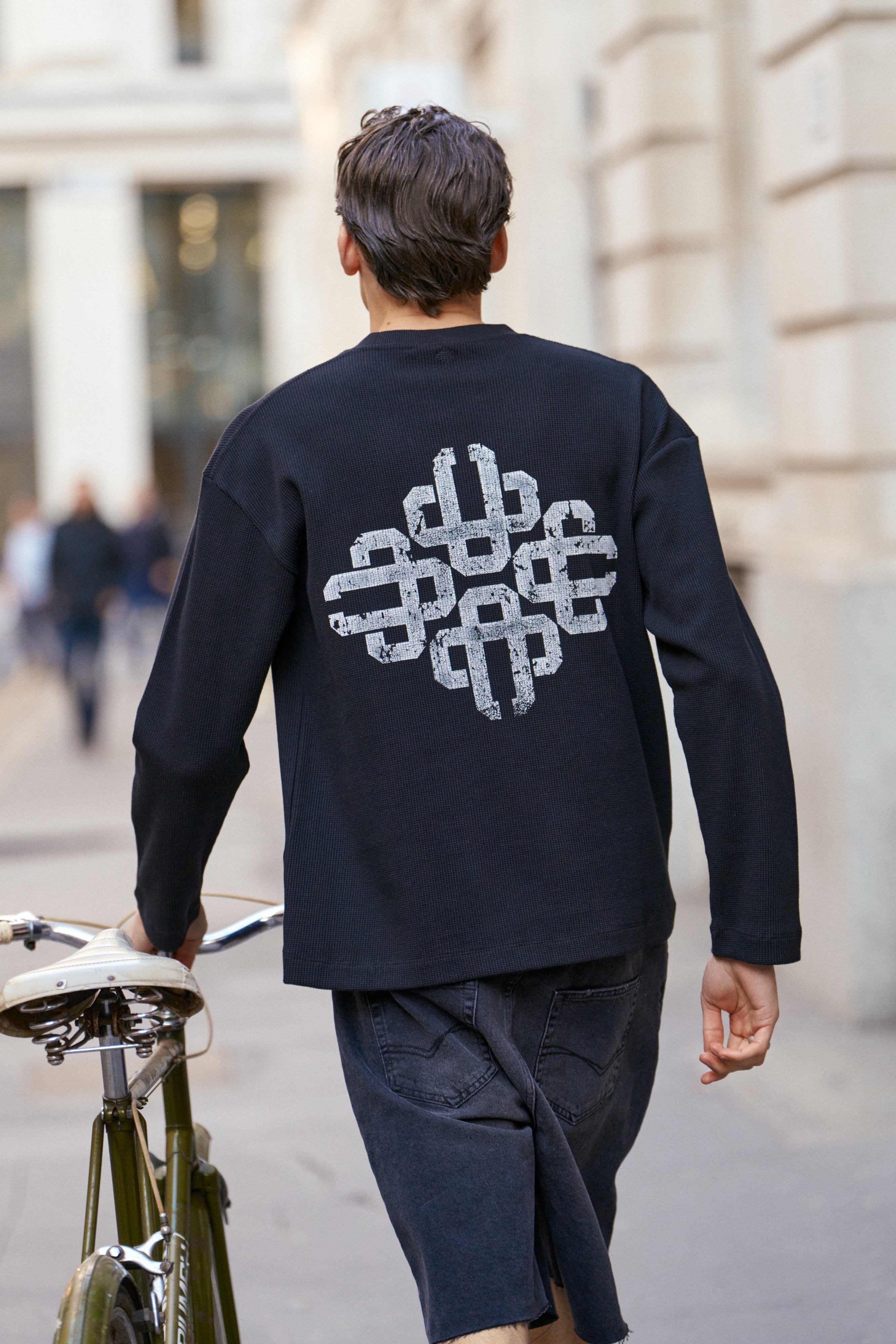 Person wearing a black t-shirt with a white geometric design, walking with a bicycle on a city street.